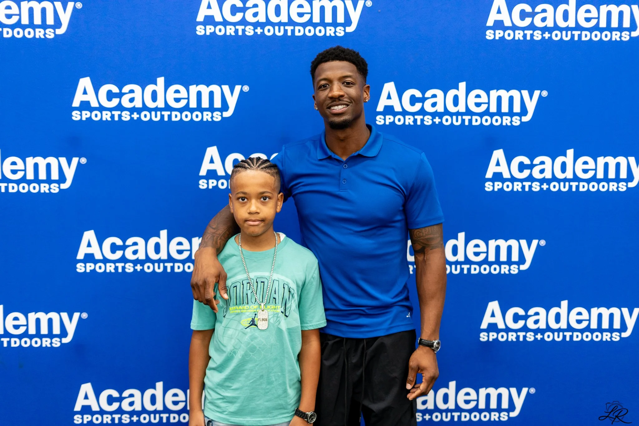 A young boy and a man standing in front of a blue backdrop with white text that reads 'Academy Sports + Outdoors.' The man is wearing a blue polo shirt and black shorts, and the boy is wearing a light green T-shirt and a dog tag necklace.