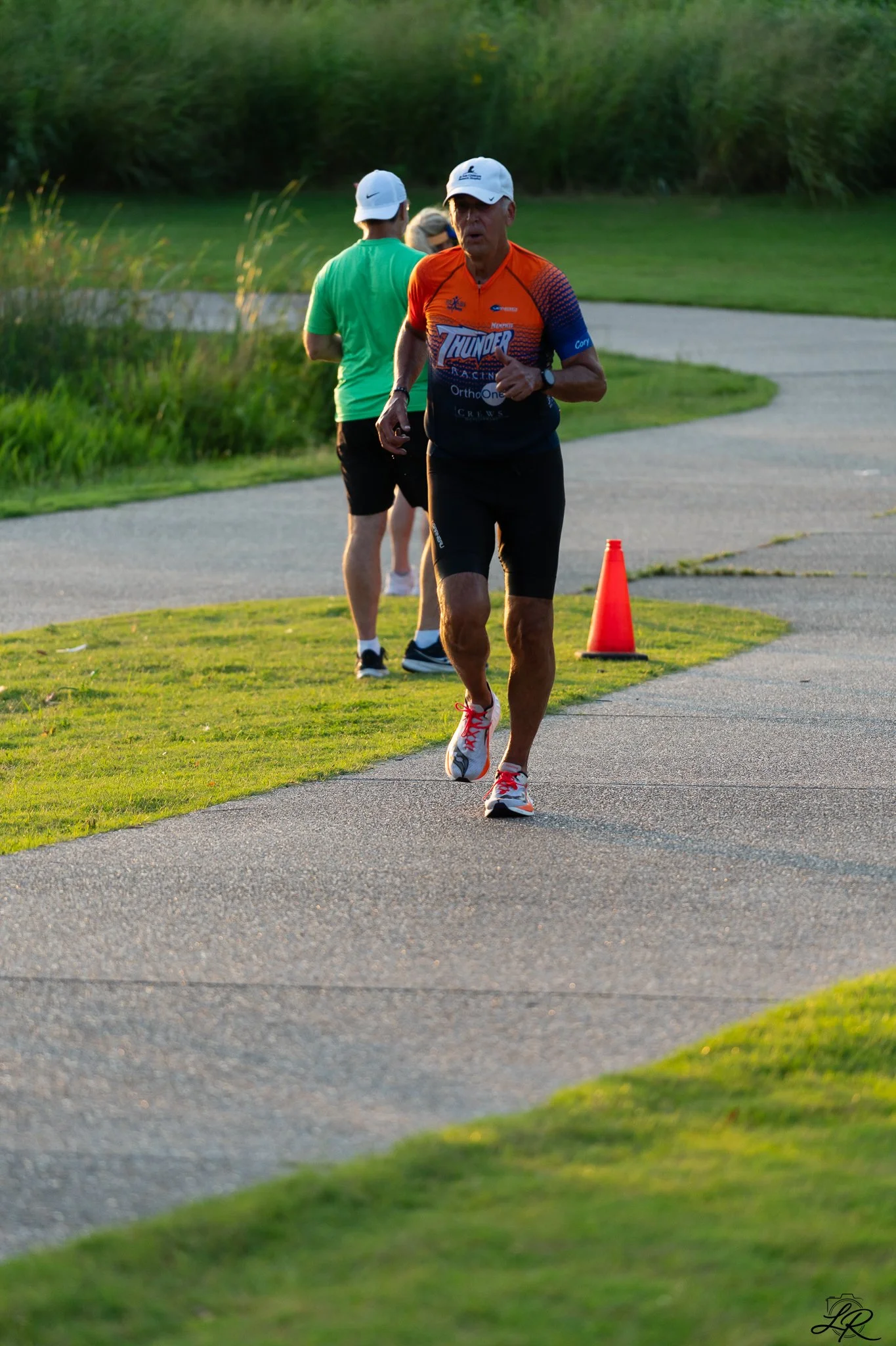 A man jogging on a paved path, wearing a bright orange and blue shirt, black shorts, and a white cap, with a green grassy area on the side and a cone in the background, during sunset.