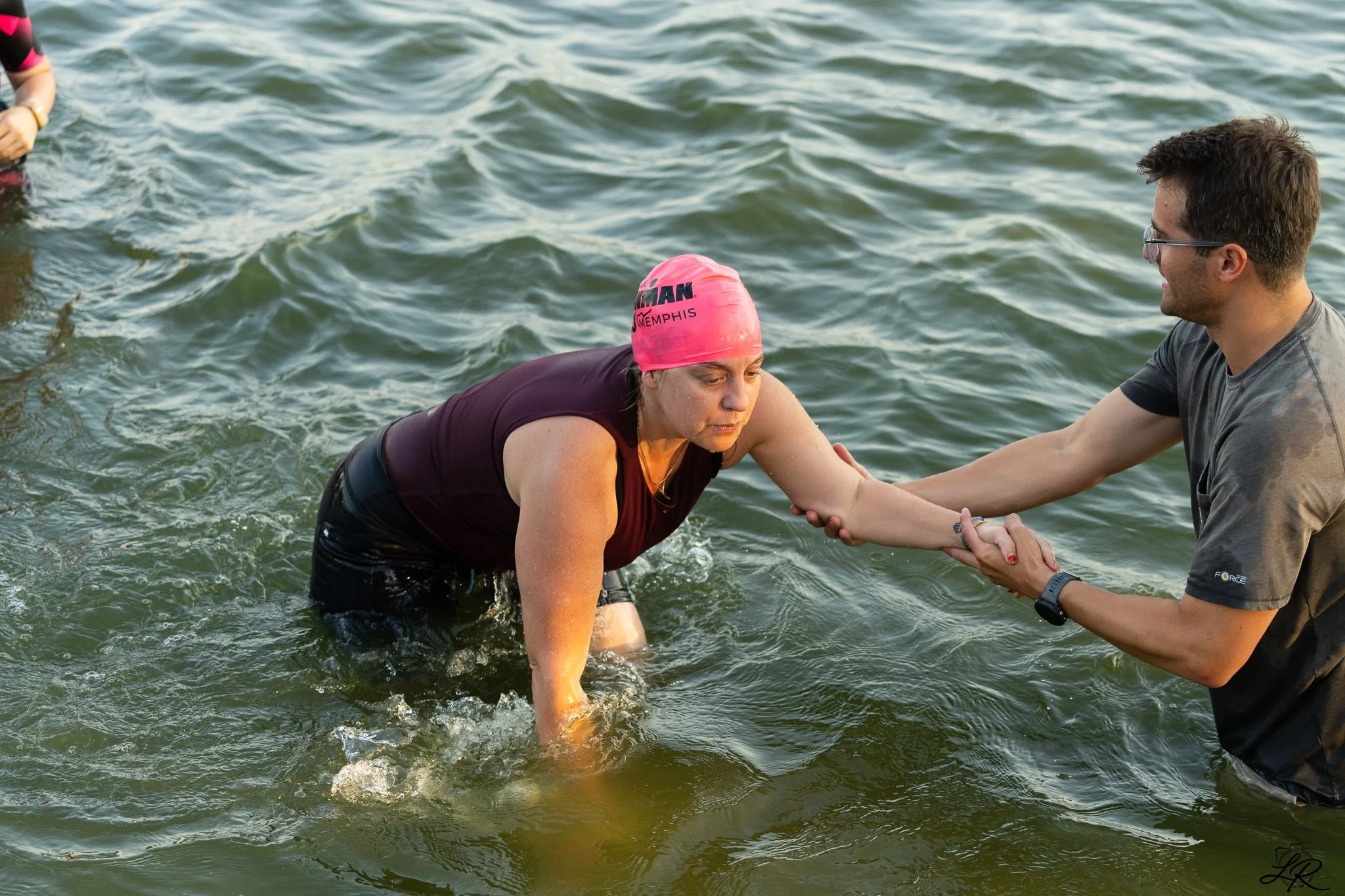 A woman wearing a pink swimming cap is being helped to stand up in water by a man holding her hands. The woman appears tired while the man smiles. They are in a body of water, possibly during a swimming or triathlon event.