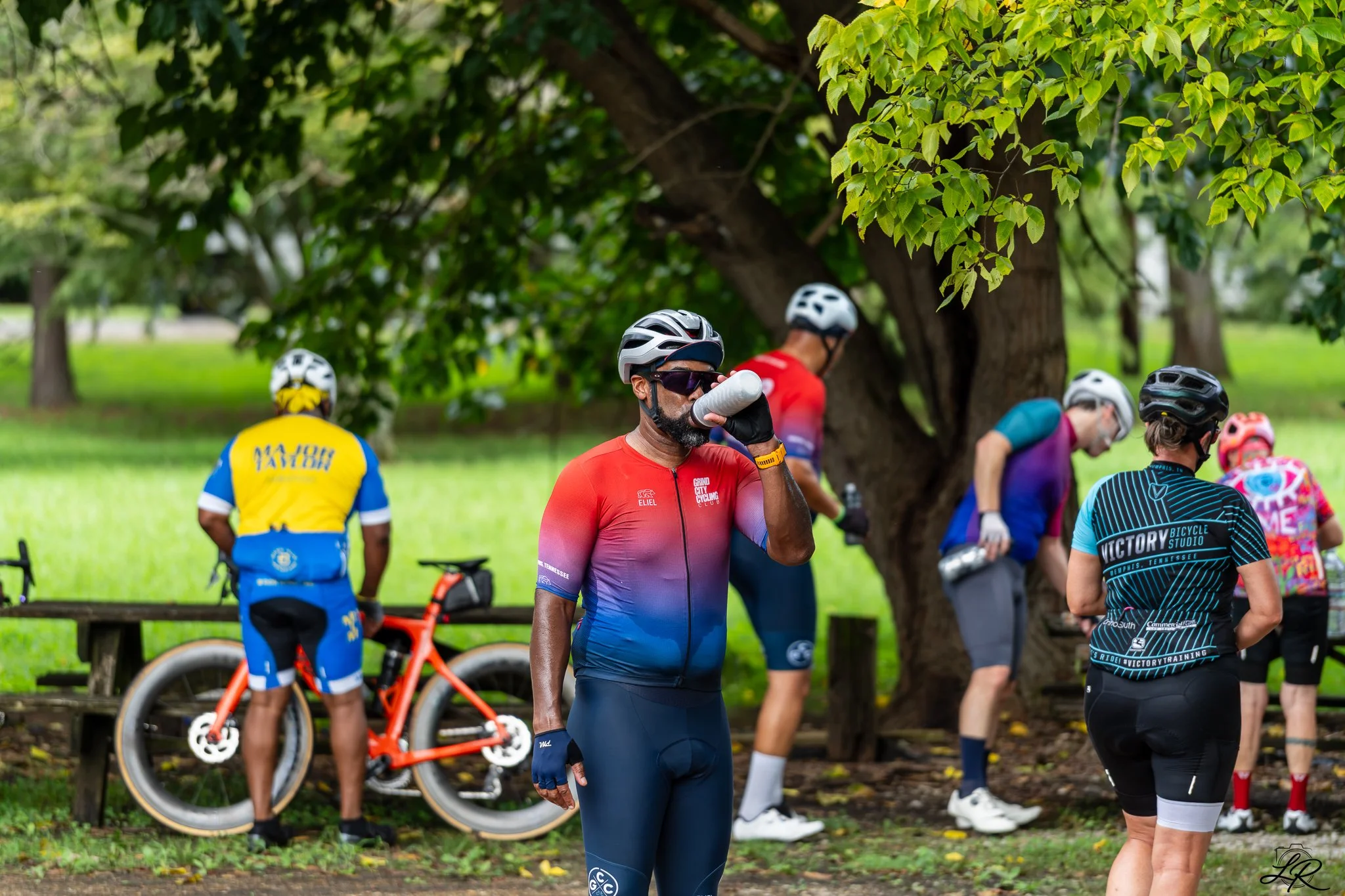 A group of cyclists resting under a large tree in a park. One cyclist in the foreground is drinking water, wearing a red cycling jersey, sunglasses, and a helmet. Other cyclists stand nearby with their bikes, including a bright orange bike, and are w