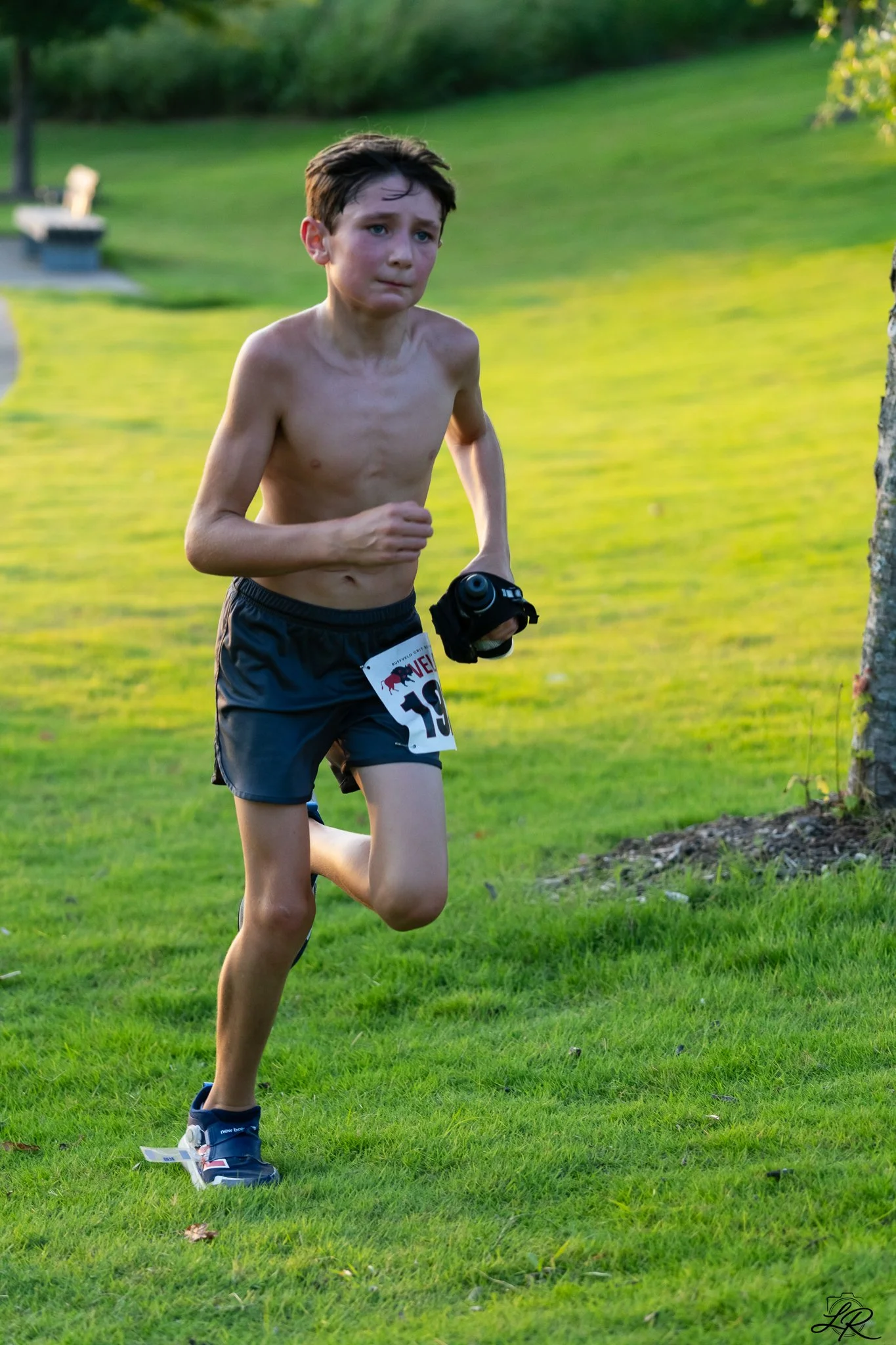 A young boy running outdoors in a park. He is shirtless, wearing black shorts with a race bib, blue running shoes, and holding a small camera. The background features green grass and trees.