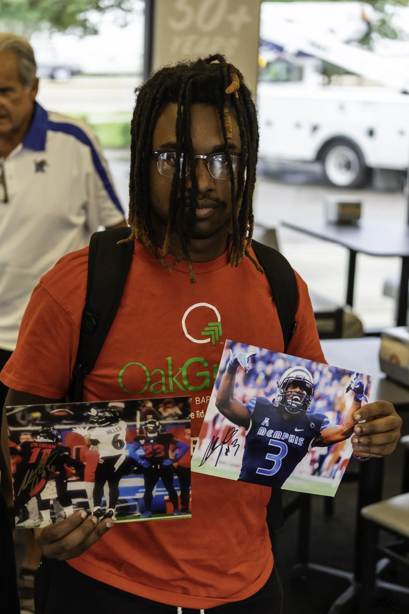 A man with dreadlocks and glasses wearing a red t-shirt is holding signed photos of football players, indoors with a window and another person in the background.