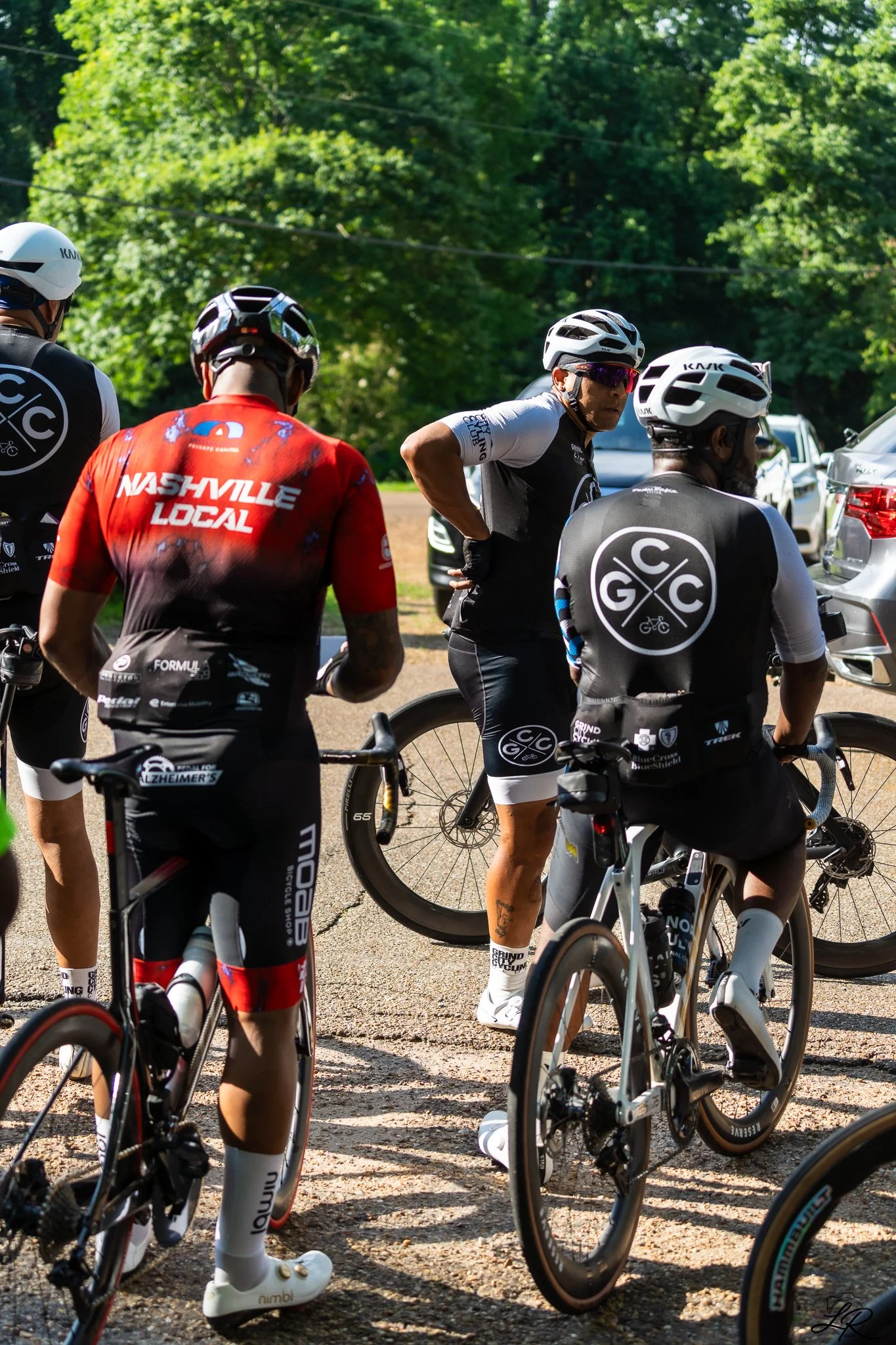 Group of cyclists gathered outdoors on a sunny day with green trees in the background, wearing cycling gear and helmets.