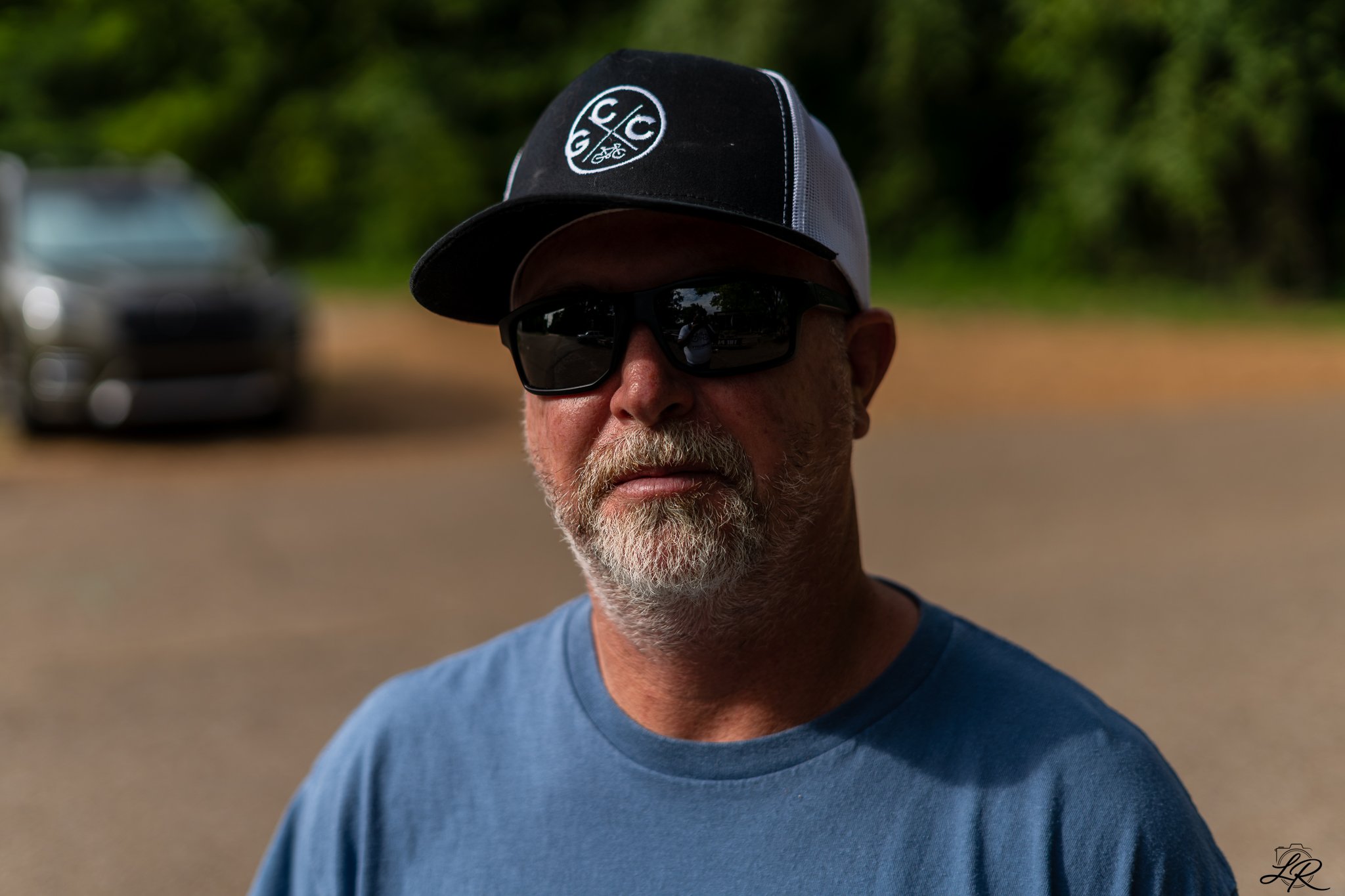 Close-up of a middle-aged man wearing black sunglasses, a blue T-shirt, a baseball cap with a logo, and a gray beard. The background shows a blurred outdoor setting with trees and a parked vehicle.
