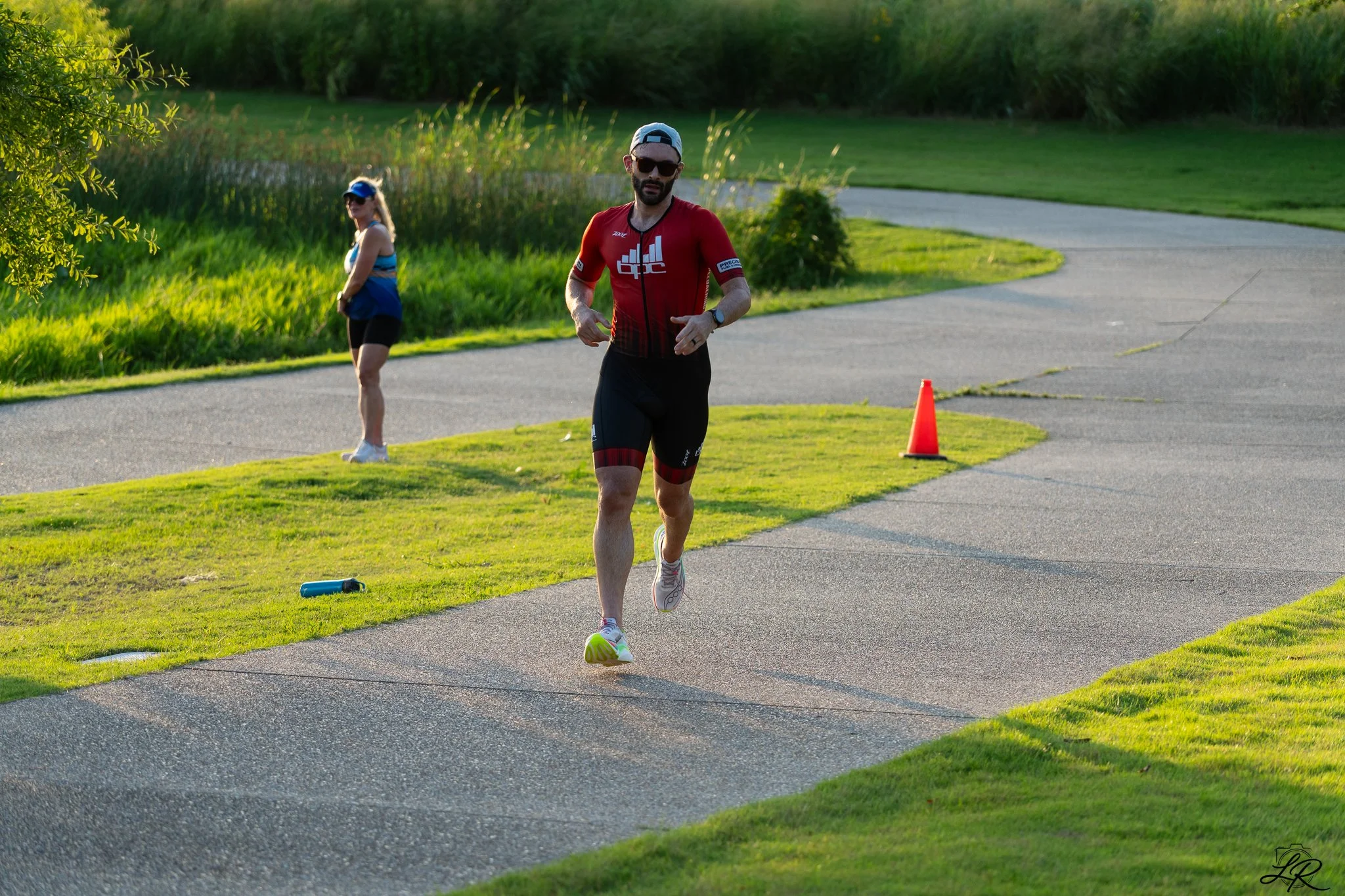 A man running outdoors on a paved path with greenery around, wearing a red athletic shirt, shorts, sunglasses, and a cap, with a woman in the background standing near the grass.