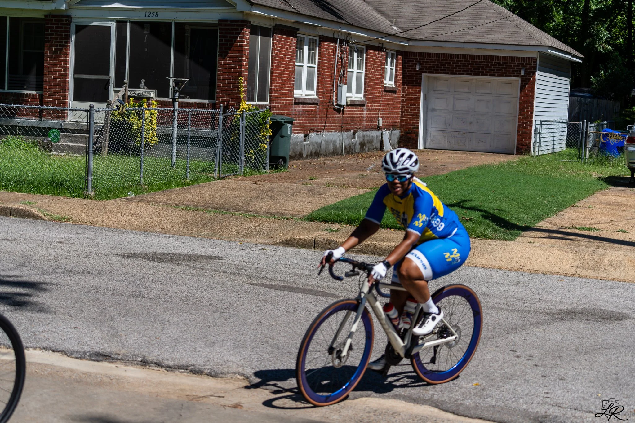 A person wearing a yellow and blue cycling jersey, black helmet, and sunglasses riding a white bike on a residential street with a brick house and green lawn in the background.