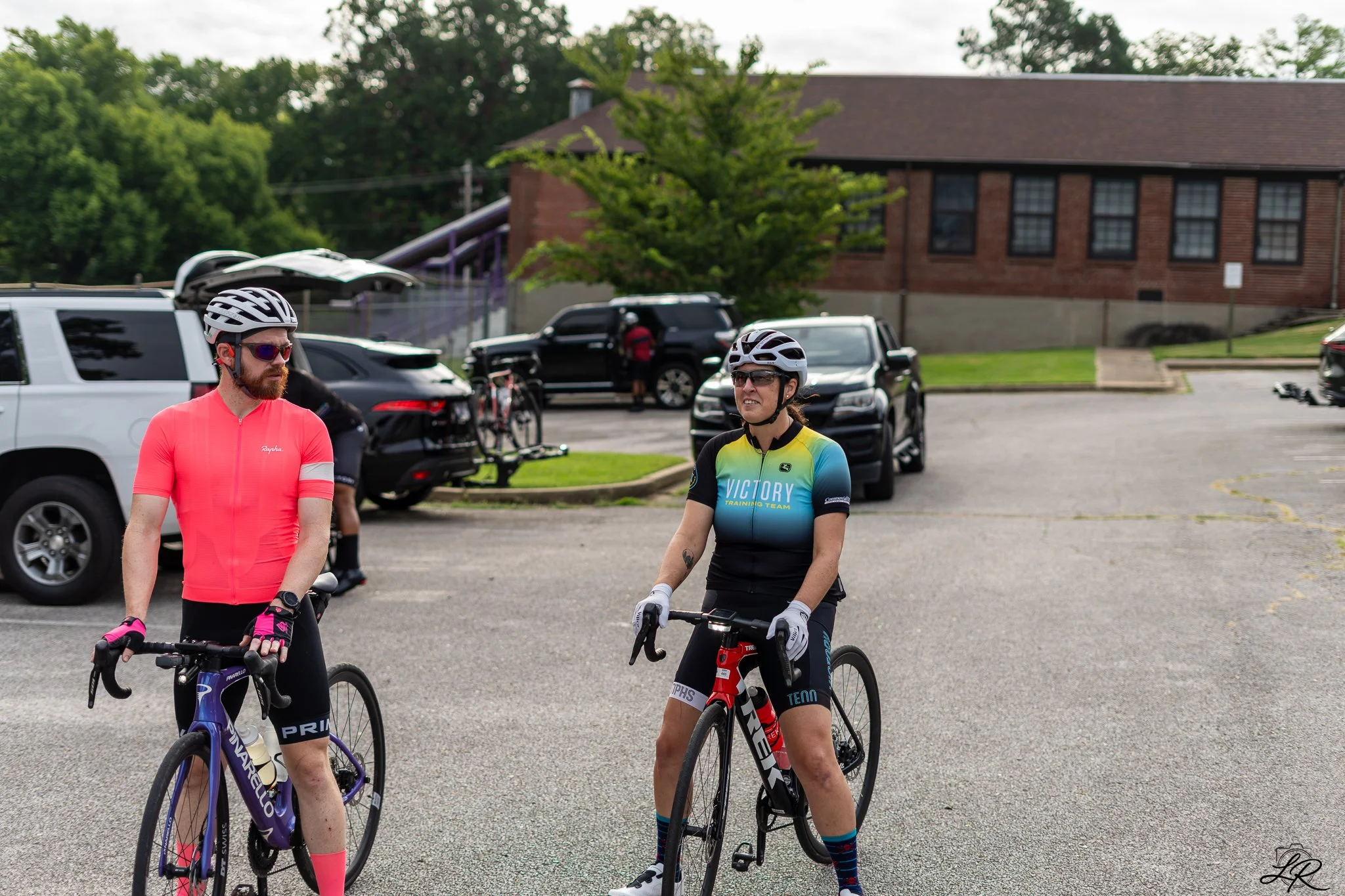 Two cyclists with helmets and sports gear standing with their bikes in a parking lot, with cars and a brick building with black window frames in the background.