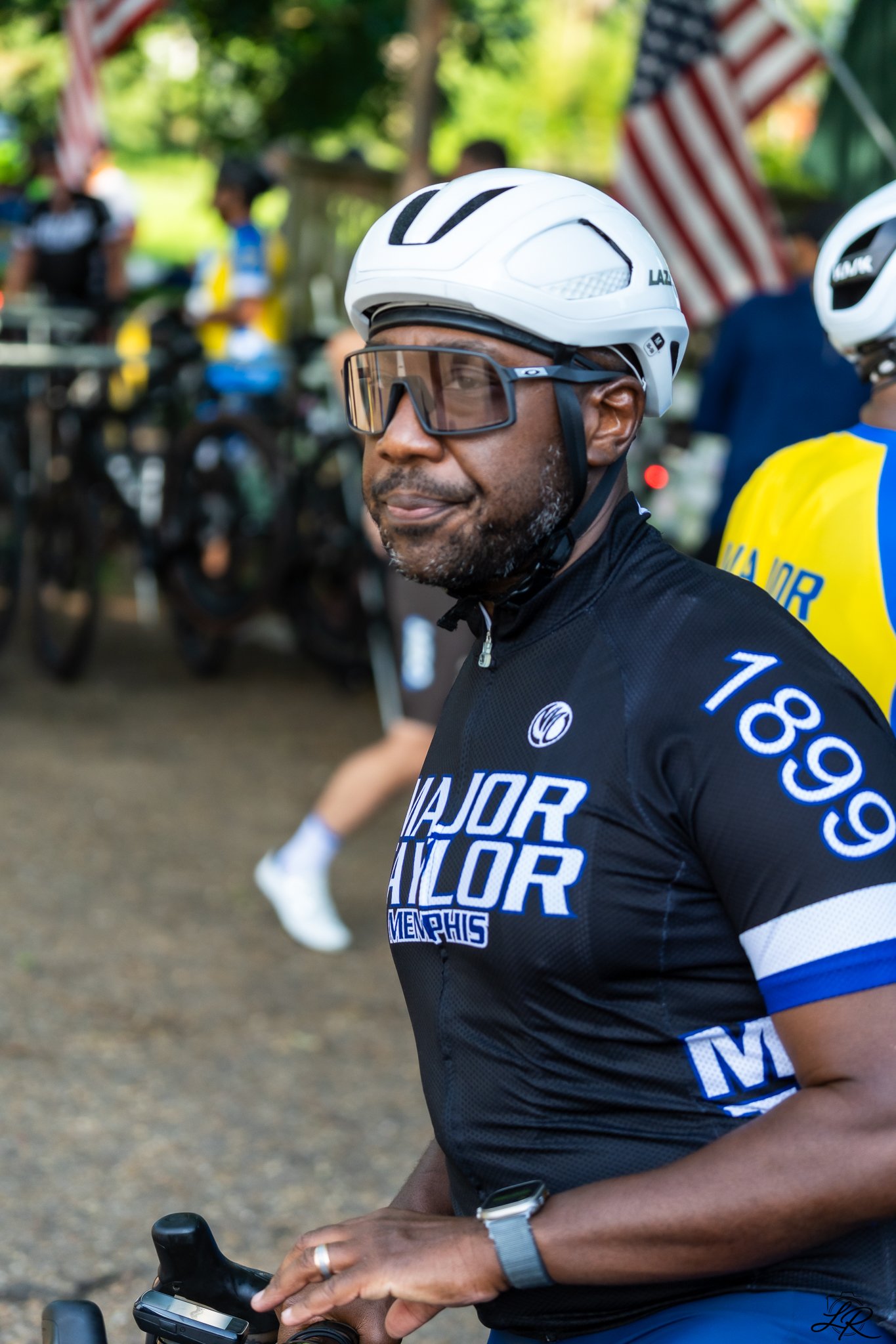 A man wearing a white bike helmet and sunglasses is riding a bicycle, holding the handlebar with both hands. He is dressed in a black and blue cycling jersey with logos and text. The background includes other cyclists, American flags, and a green out