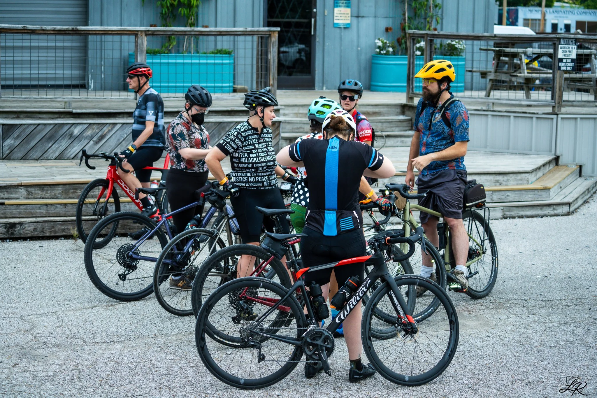 Group of cyclists wearing helmets and sunglasses gathered outdoors near wooden stairs, with some on mountain and road bikes, engaged in conversation.