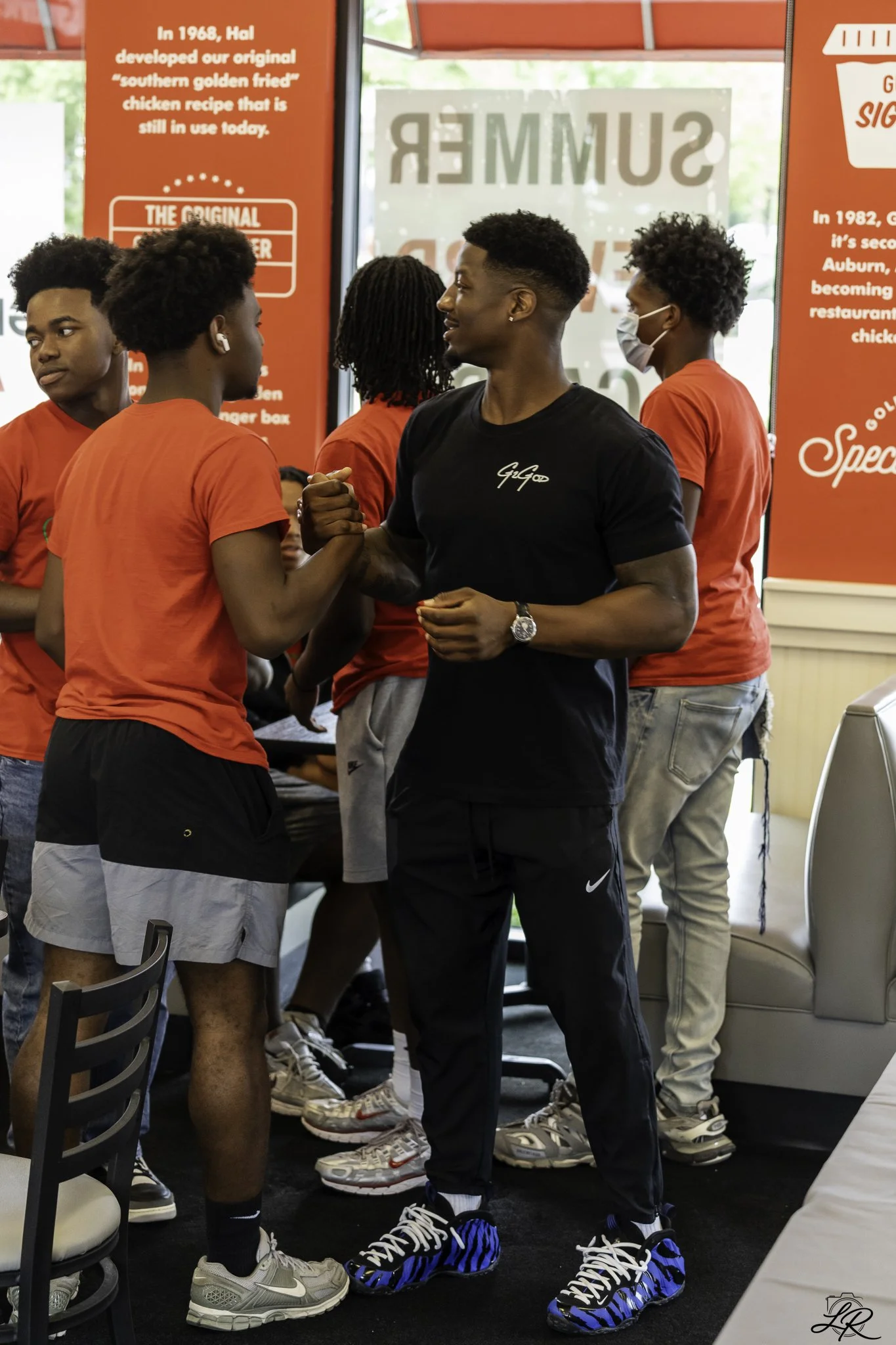 A group of young men at a fast-food restaurant, some wearing red shirts, one with a face mask, shaking hands, and some wearing sneakers with black and blue designs. Red signs with white text are in the background.