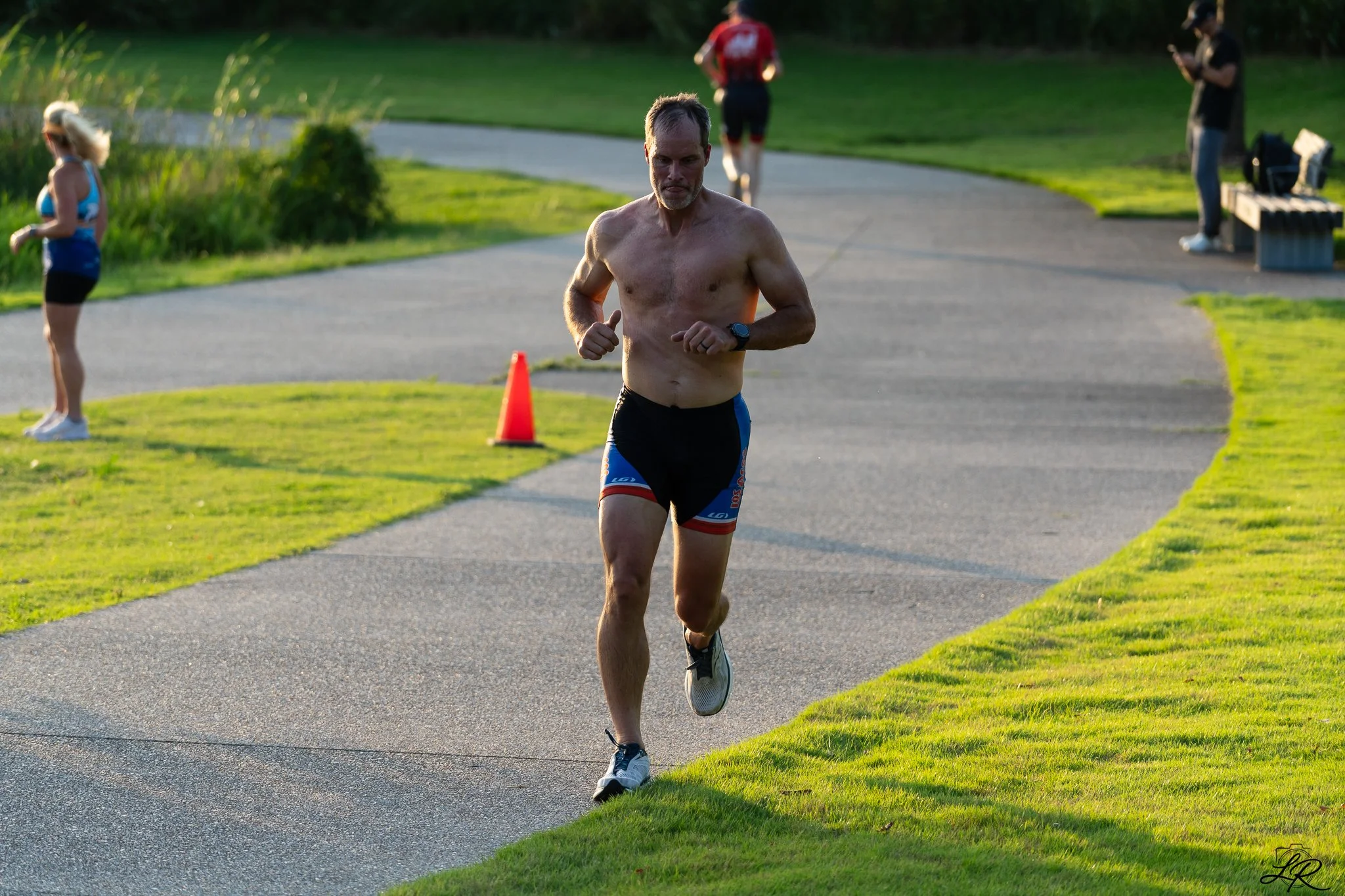 A man running shirtless along a paved path in a park with green grass and trees, with a woman and two other people in the background.