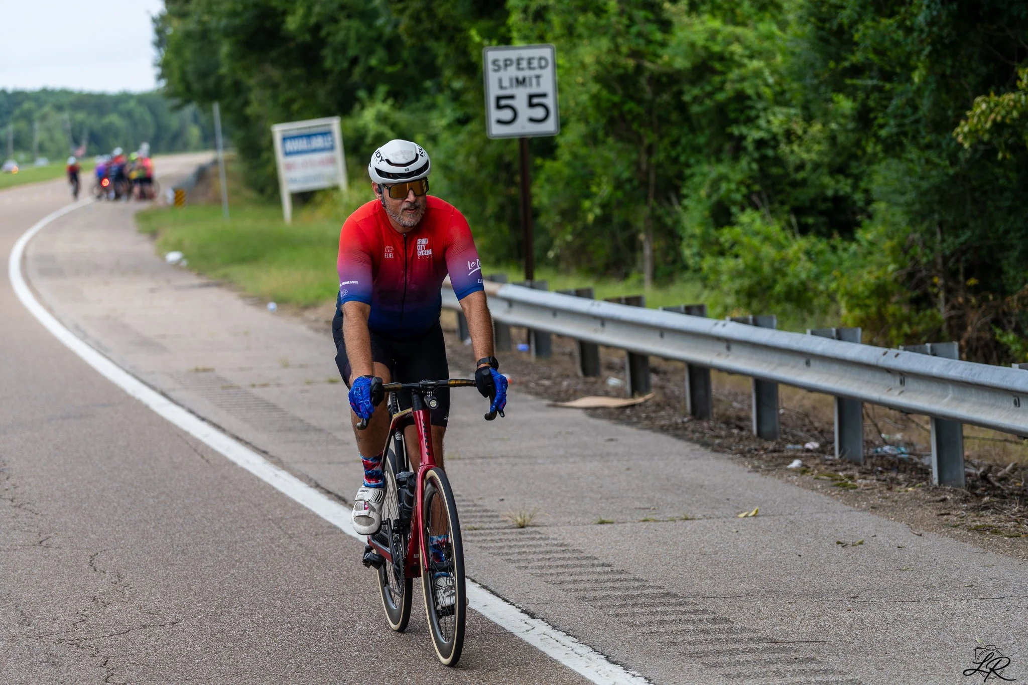 A man wearing a white helmet, sunglasses, and a red cycling jersey rides a road bike along a paved road with a speed limit sign of 55 mph, surrounded by green trees and bushes.