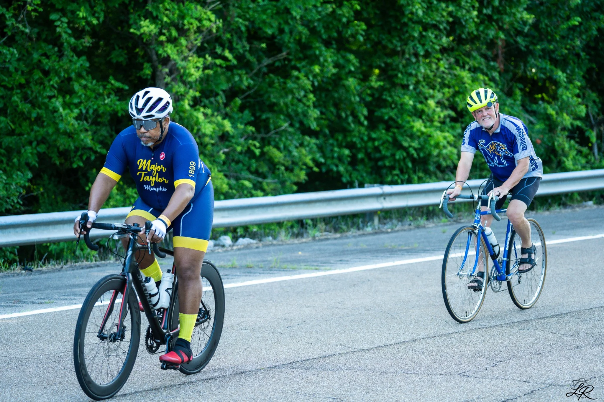 Two men riding bicycles on a road with a metal guardrail and lush green trees in the background. The man on the left is wearing a white helmet, sunglasses, and a blue cycling jersey with yellow accents. The man on the right is wearing a yellow helmet