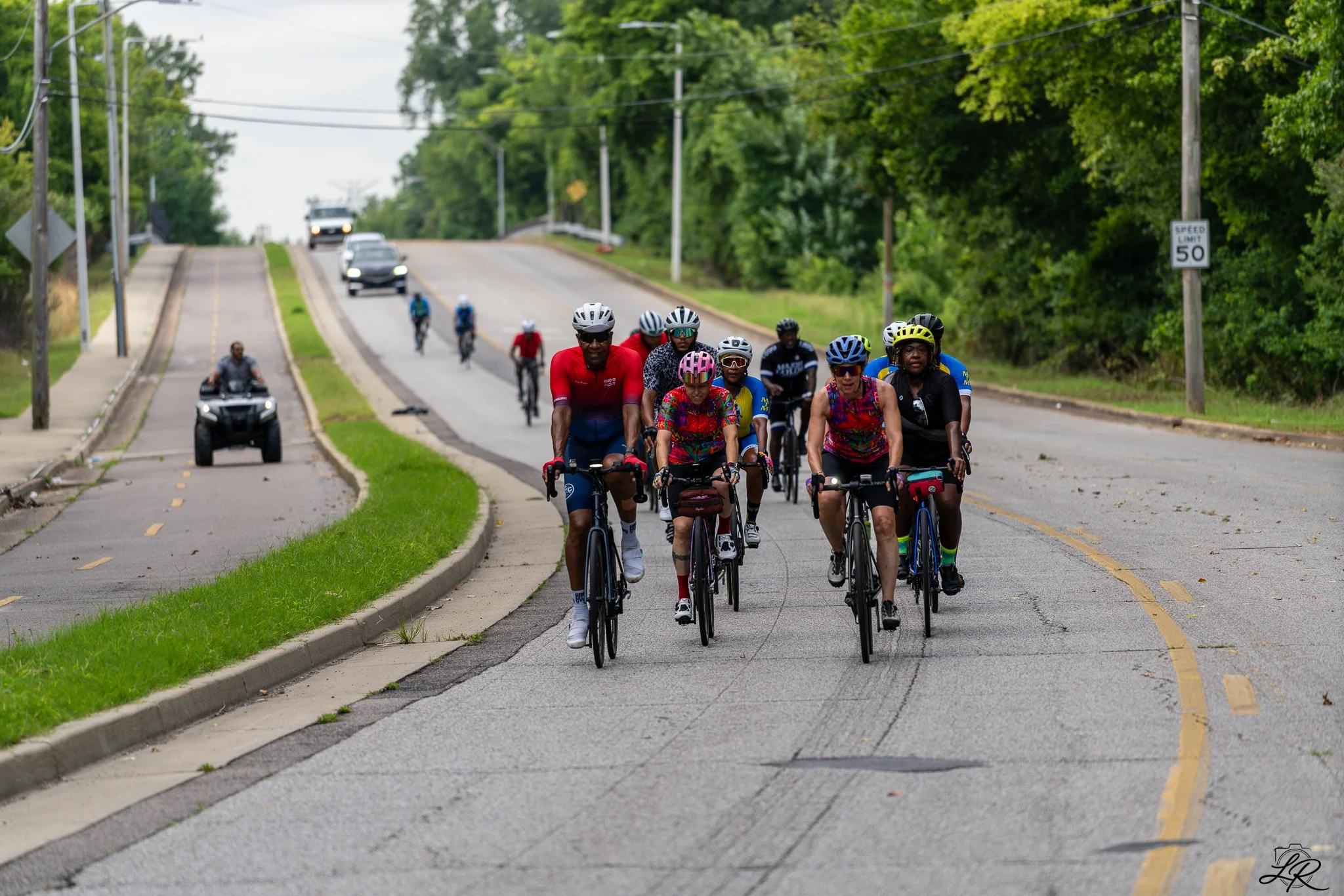 A group of cyclists riding on a city street with cars and motorcycles nearby, surrounded by green trees and power lines.
