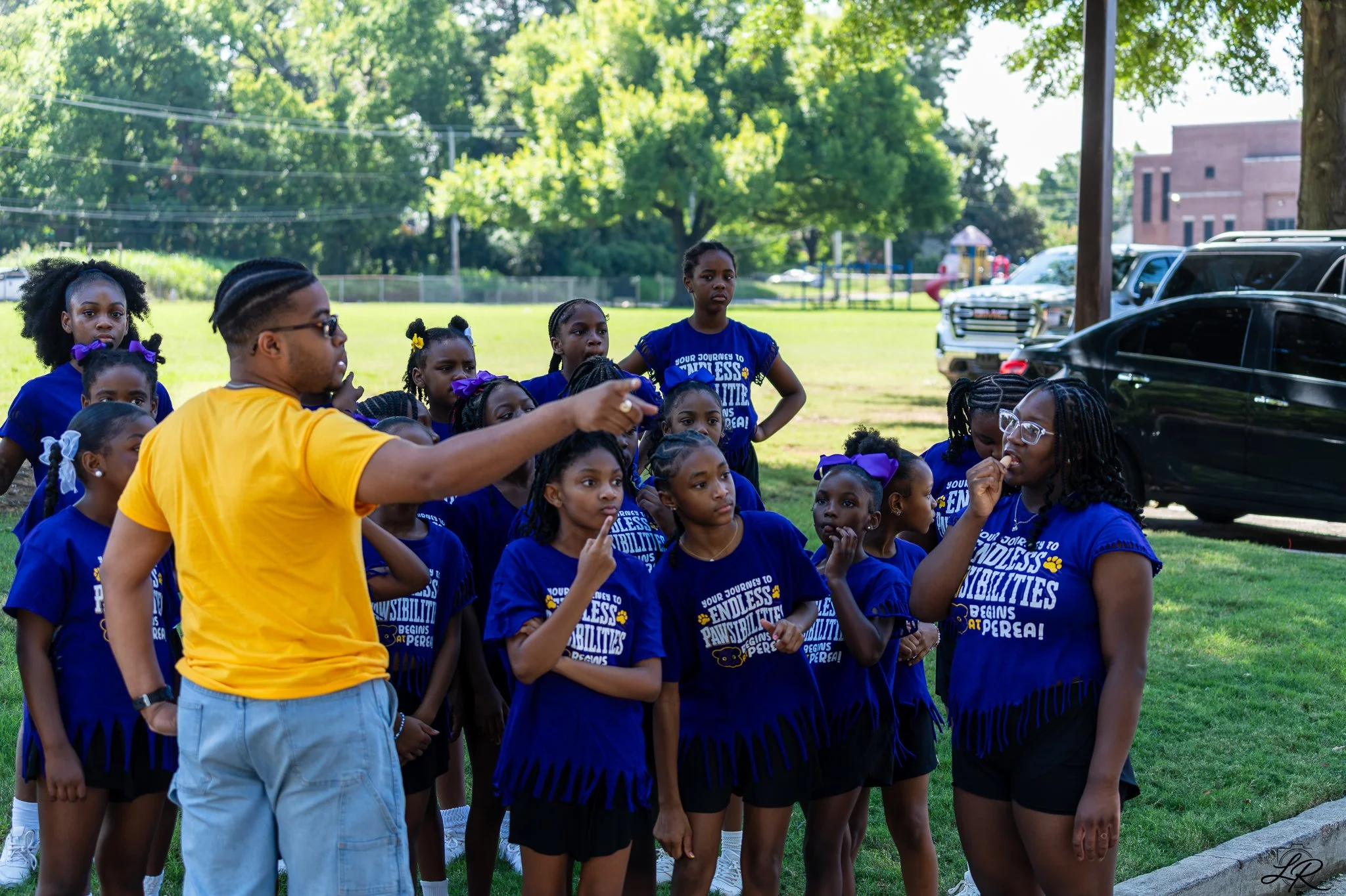 A group of young girls in blue shirts and purple bows standing outdoors on a grassy area, listening to a man in a yellow shirt who is speaking to them. There are trees, parked cars, and a playground in the background.