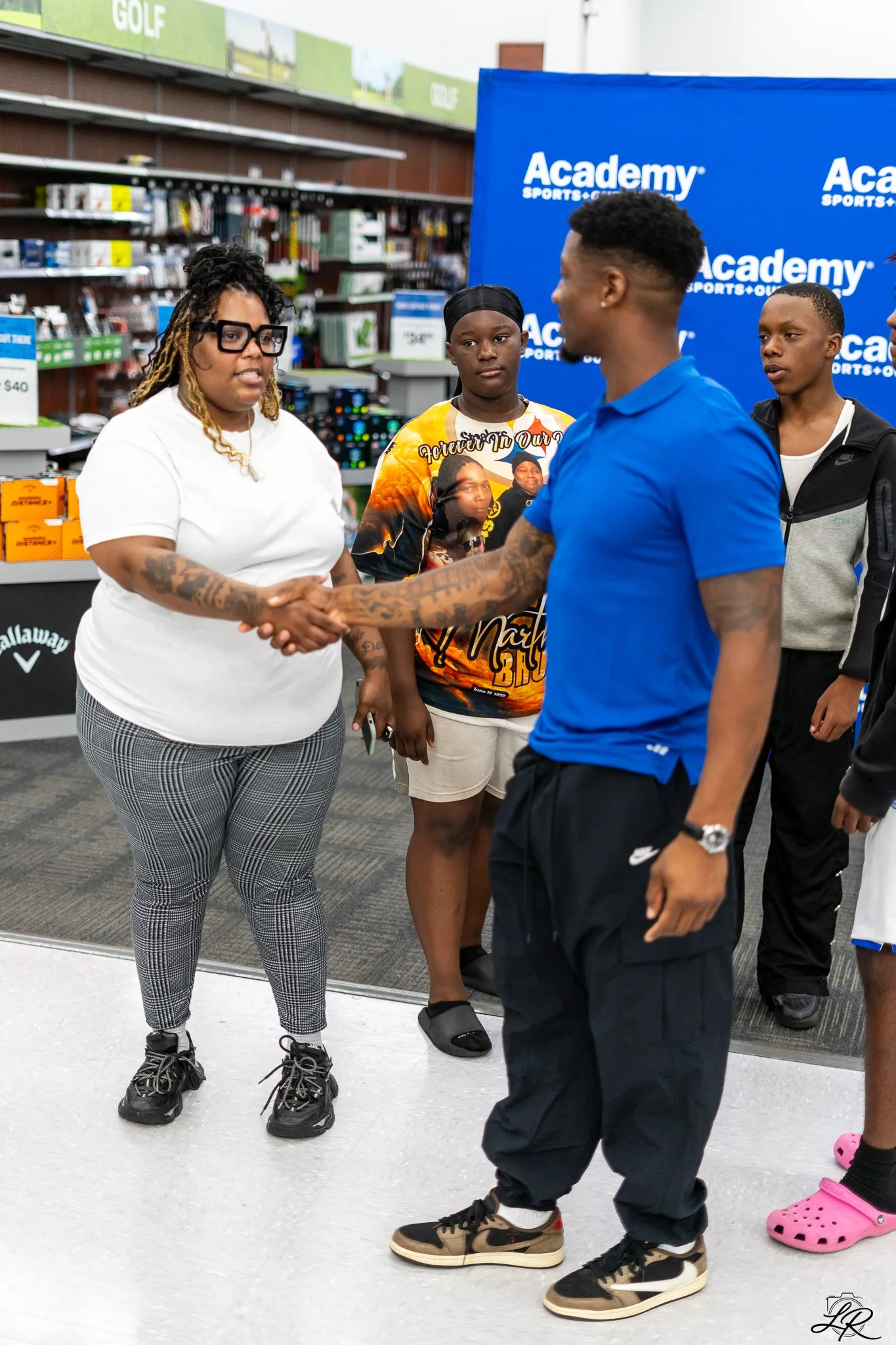 A woman with glasses shaking hands with a man wearing a blue shirt in an outdoor store setting. Three other young people are standing nearby, two of them observing the interaction. The background features store shelves and a blue Academy Sports + Out