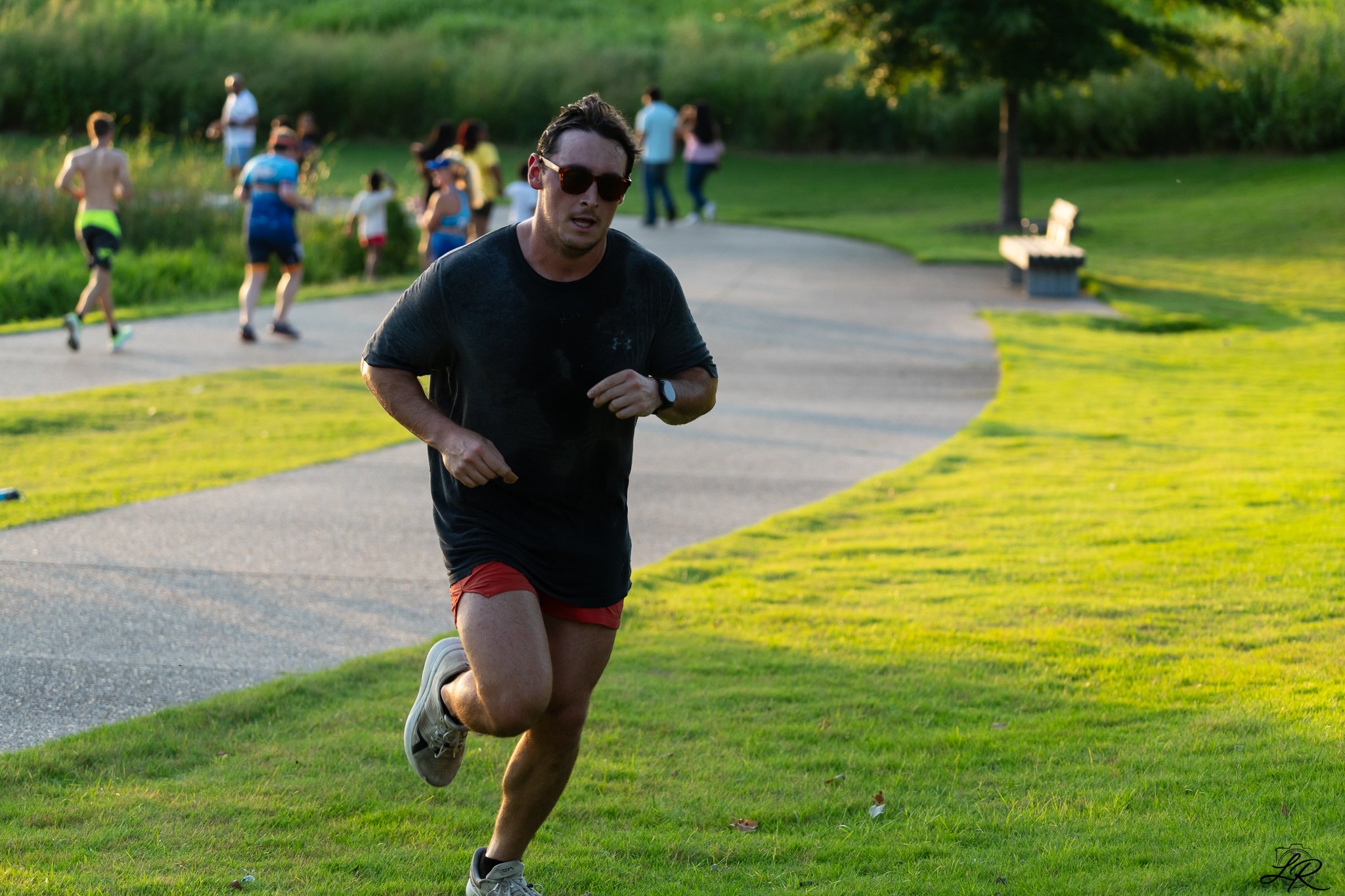 A man in a black athletic shirt, red shorts, and sunglasses is running on a park pathway, with a group of people in the background near a pond, some standing and some walking. The park has green grass and trees.