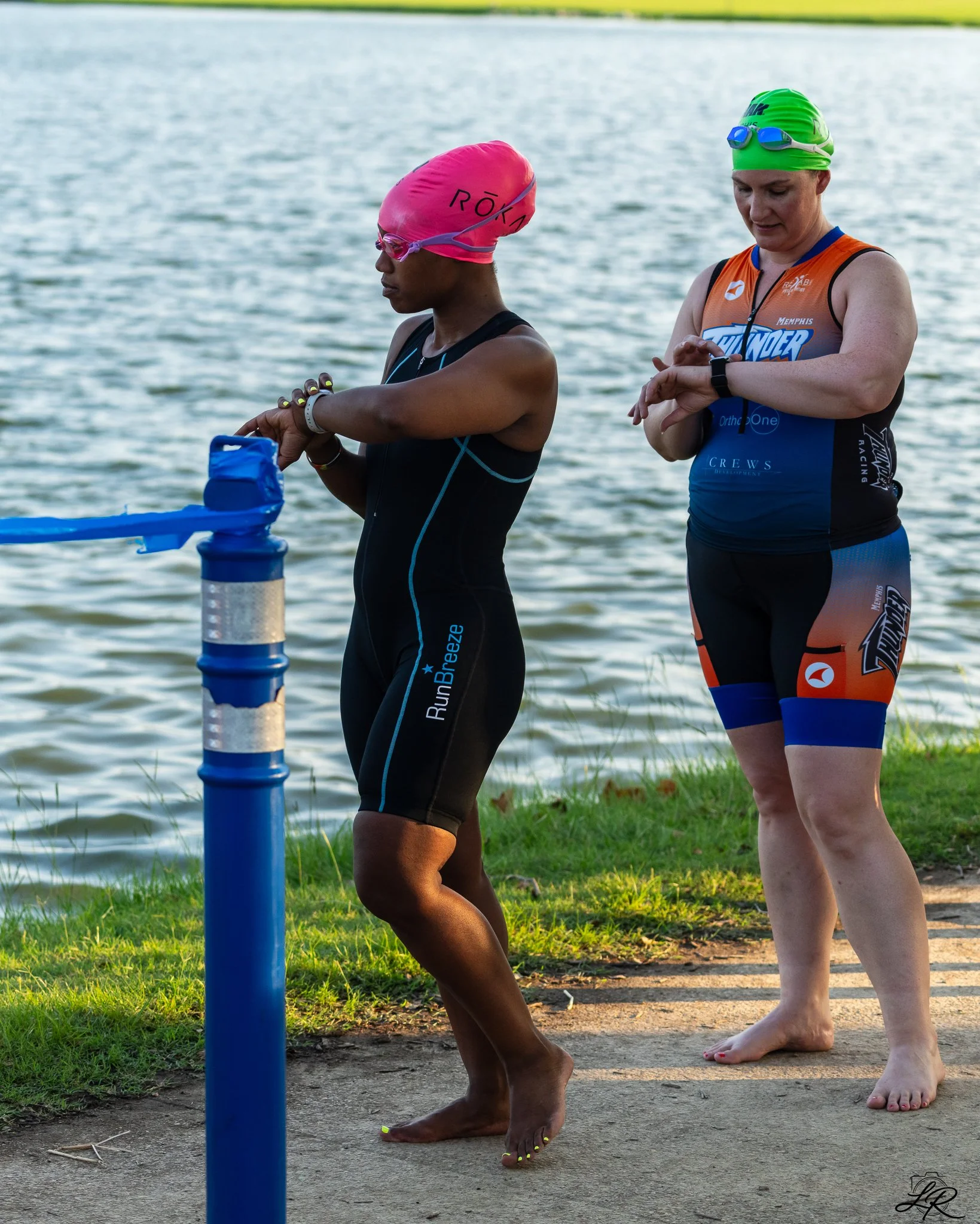 Two women standing by a body of water checking their watches. One wears a bright pink swim cap and a black wetsuit, and the other wears a bright green swim cap and an orange and black triathlon suit. They are preparing for a swim race.