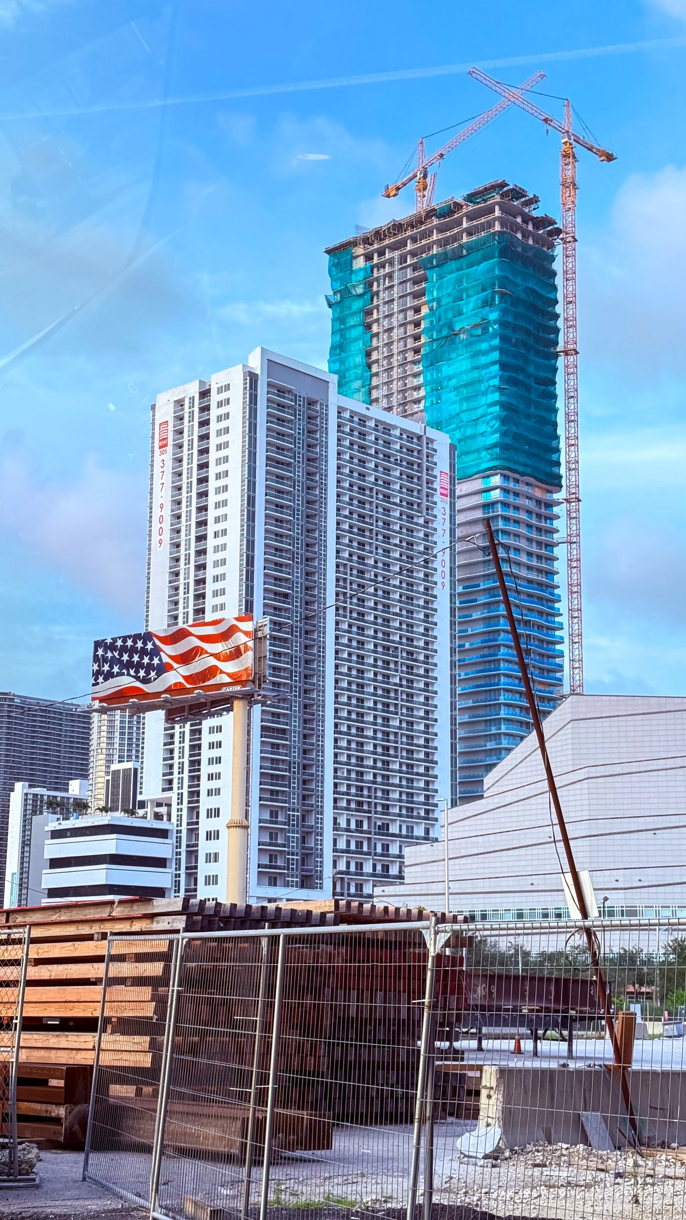 Construction site with high-rise buildings under construction, a crane at the top, a digital billboard displaying an American flag, fencing around the site, and partly cloudy sky.