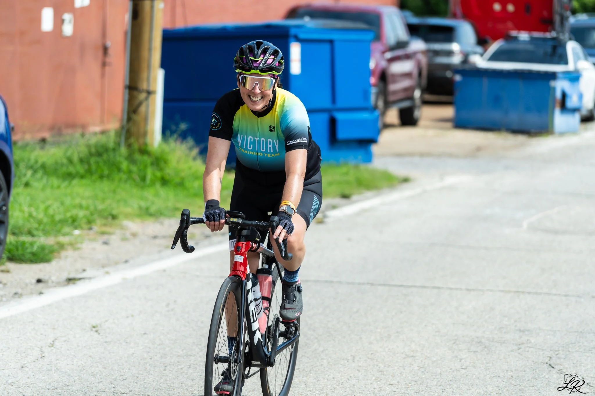 A woman riding a black and red bicycle on a paved road, wearing a black helmet, sunglasses, a yellow and blue cycling jersey, black shorts, and gloves, smiling while riding.