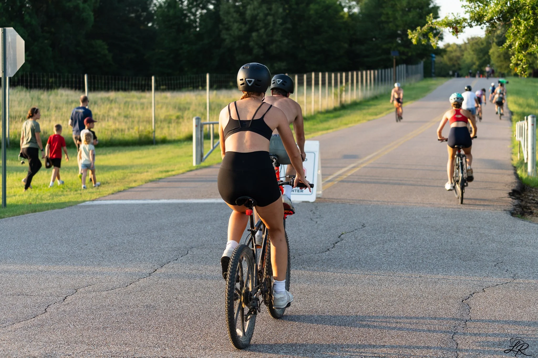 People riding bicycles on a paved trail with a grassy field and trees in the background, some wearing helmets, in the late afternoon or early evening.