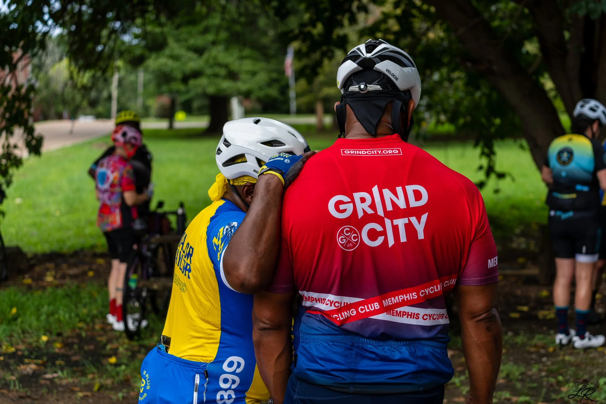 Two cyclists wearing helmets and colorful jerseys embracing each other outdoors under trees, with other people and bicycles visible in the background.