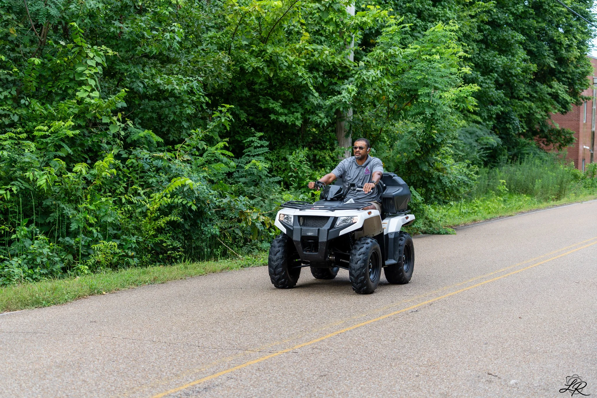 Man riding an all-terrain vehicle (ATV) on a paved road with green trees on the side.