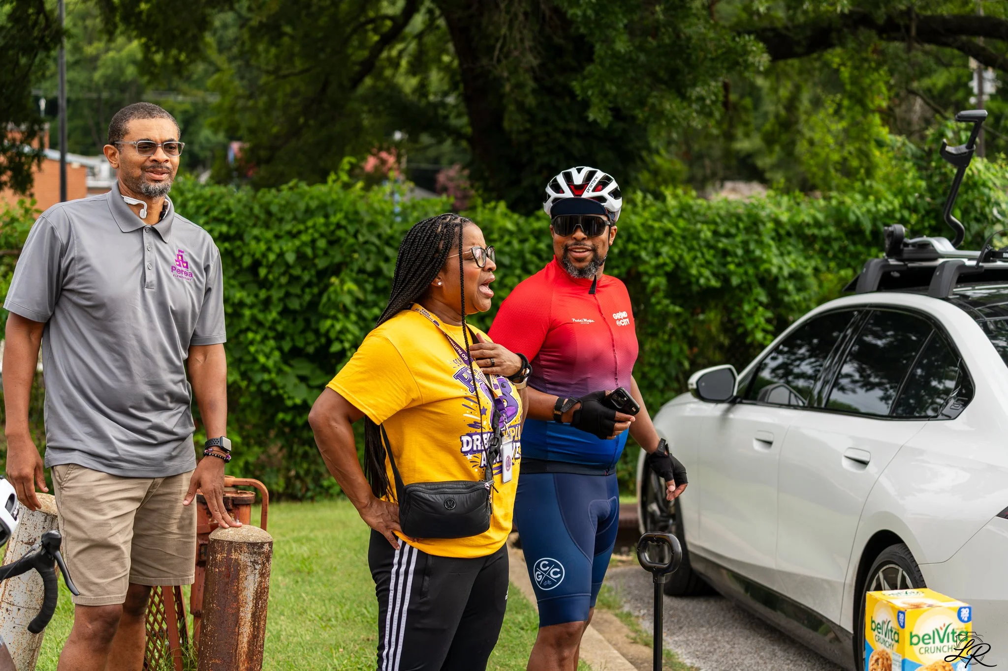 Three people standing outdoors near a white car, with a tree and green shrubbery in the background. The woman in the center, wearing a yellow shirt and glasses, is speaking to the man on the right, who is wearing a red cycling shirt, a helmet, and su