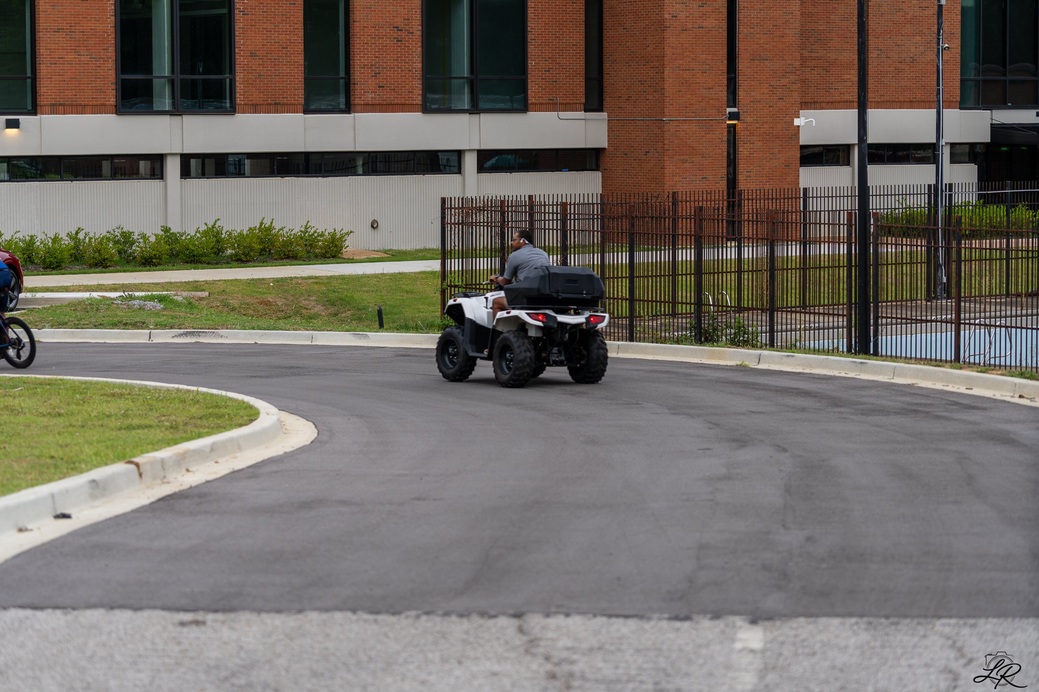 A person riding an all-terrain vehicle on a paved road in front of a fenced area with a multi-story red brick building in the background.