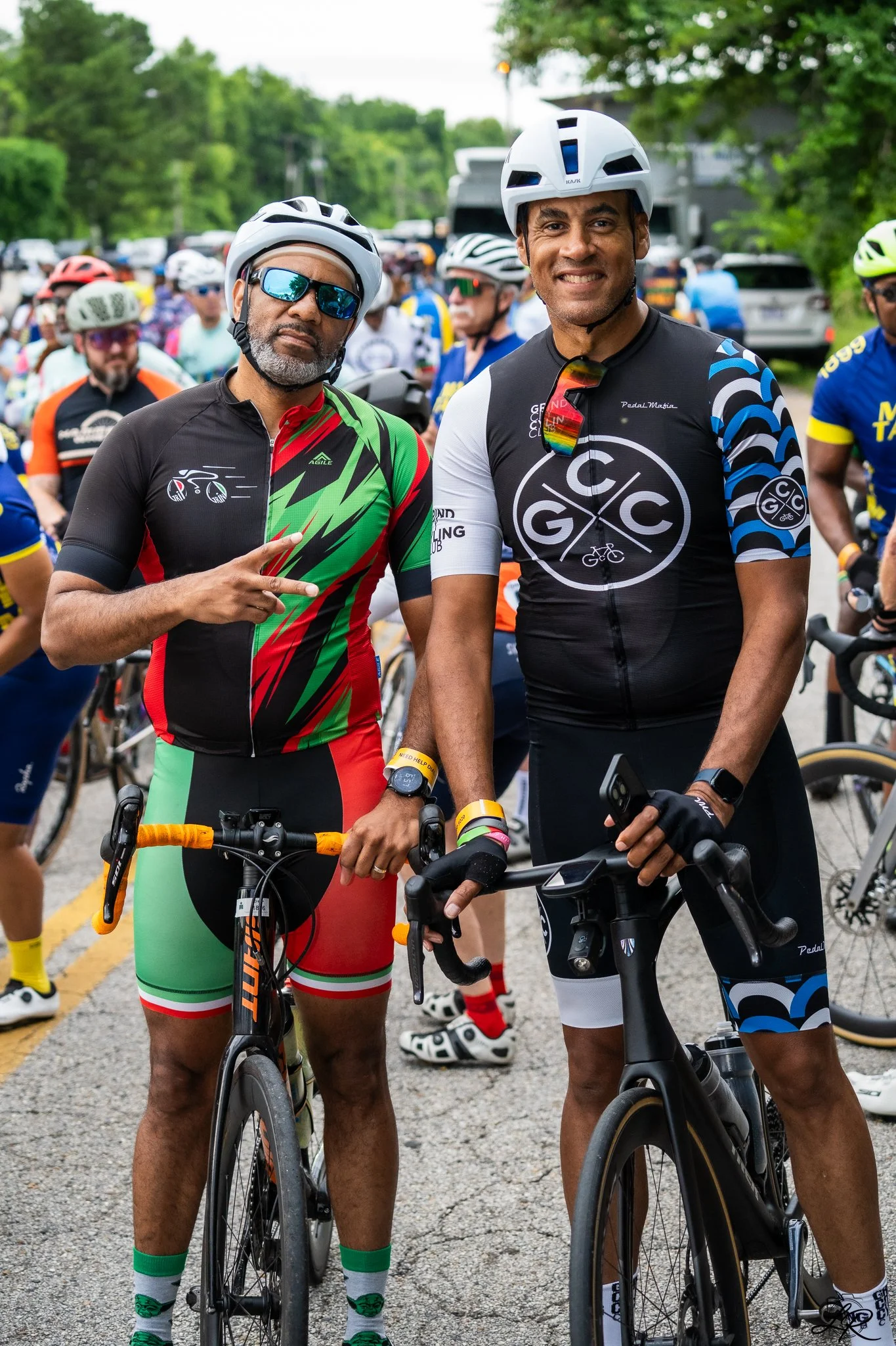Two men in cycling gear and helmets at a cycling event, standing with bikes and smiling for the camera. A crowd of cyclists is in the background on a tree-lined street.