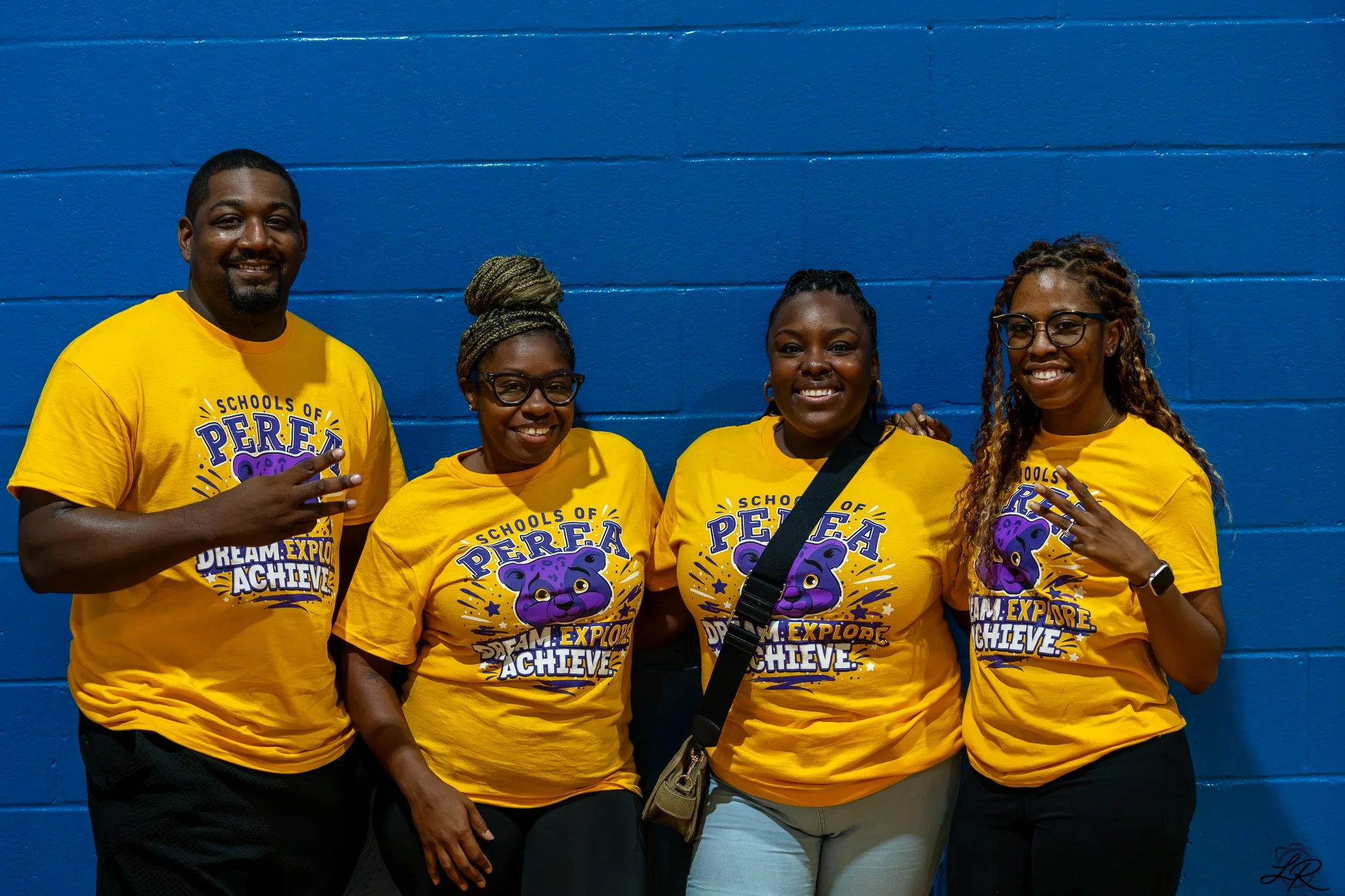 Four people wearing yellow T-shirts with a purple and black graphic of a lion and motivational text standing against a blue brick wall. They are smiling and posing for a group photo.