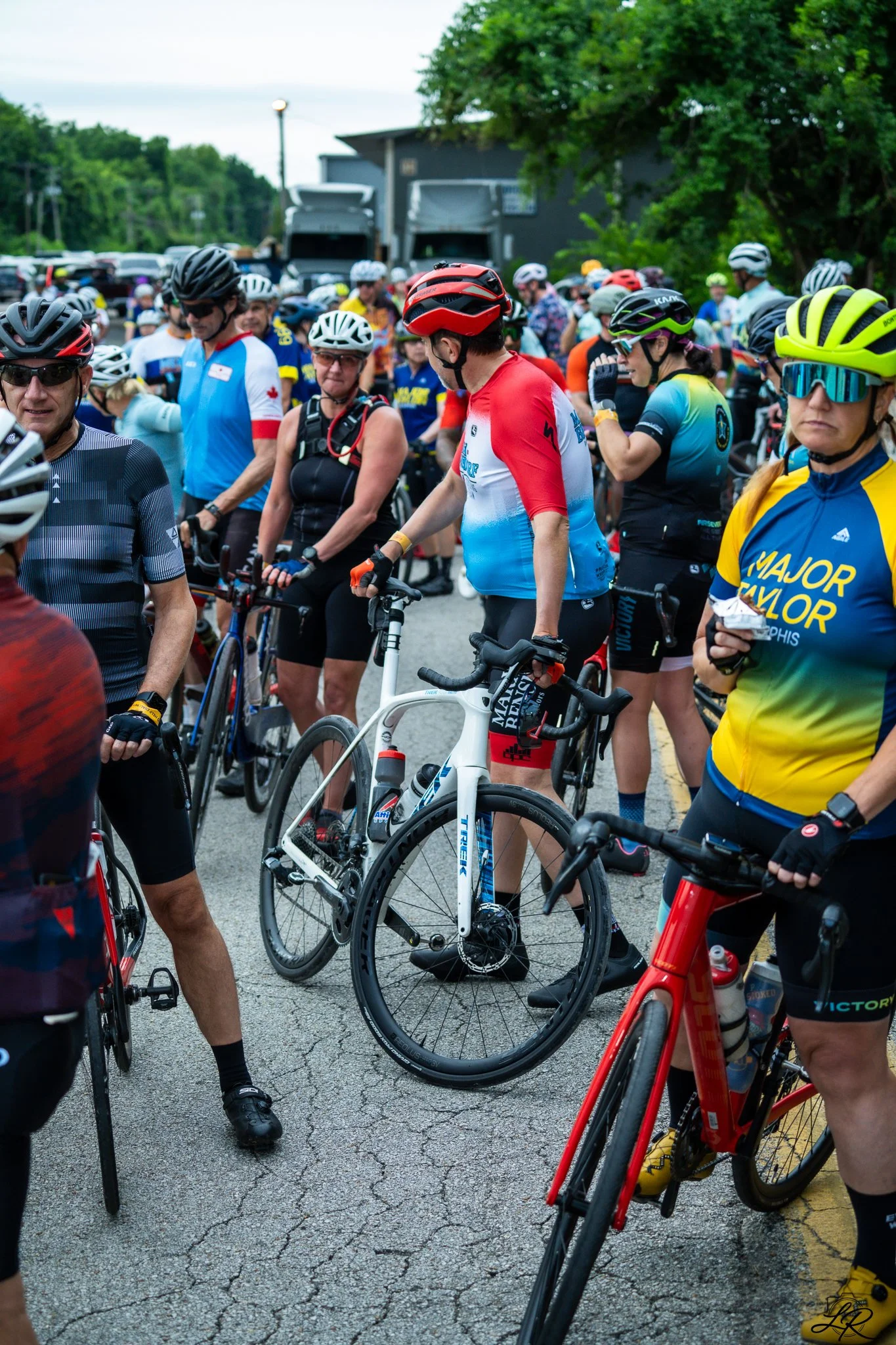 Cyclists gather at a starting line, many wearing helmets and athletic gear, with trees and trucks in the background.