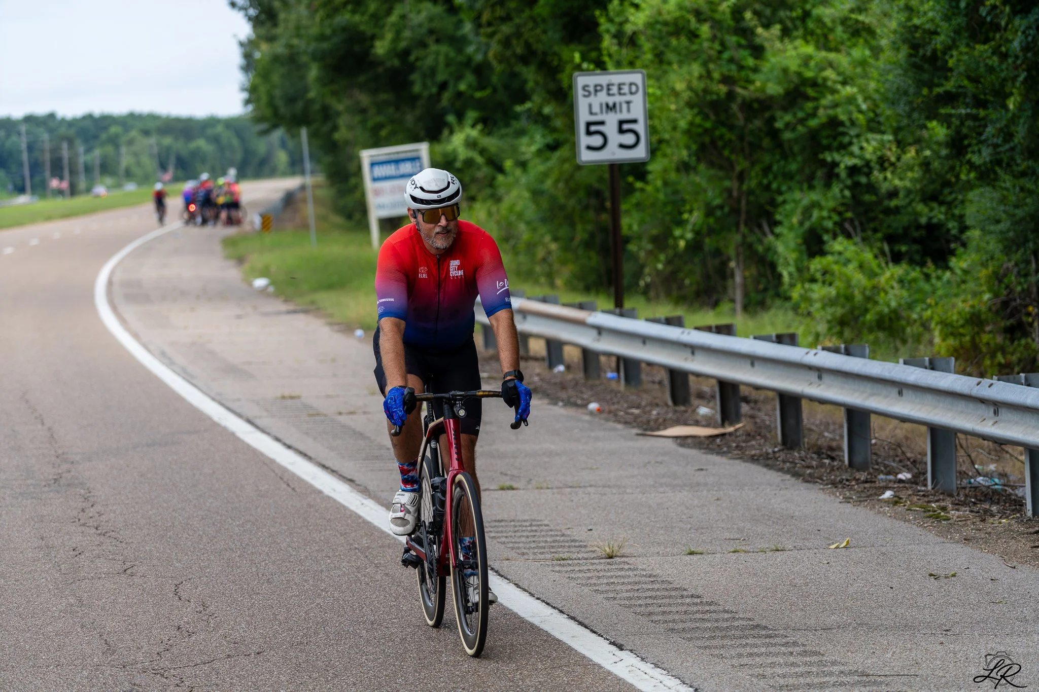 A man in a red cycling jersey and helmet rides a bicycle along a rural road with a speed limit sign of 55 mph. In the background, there are several other cyclists and lush green trees.