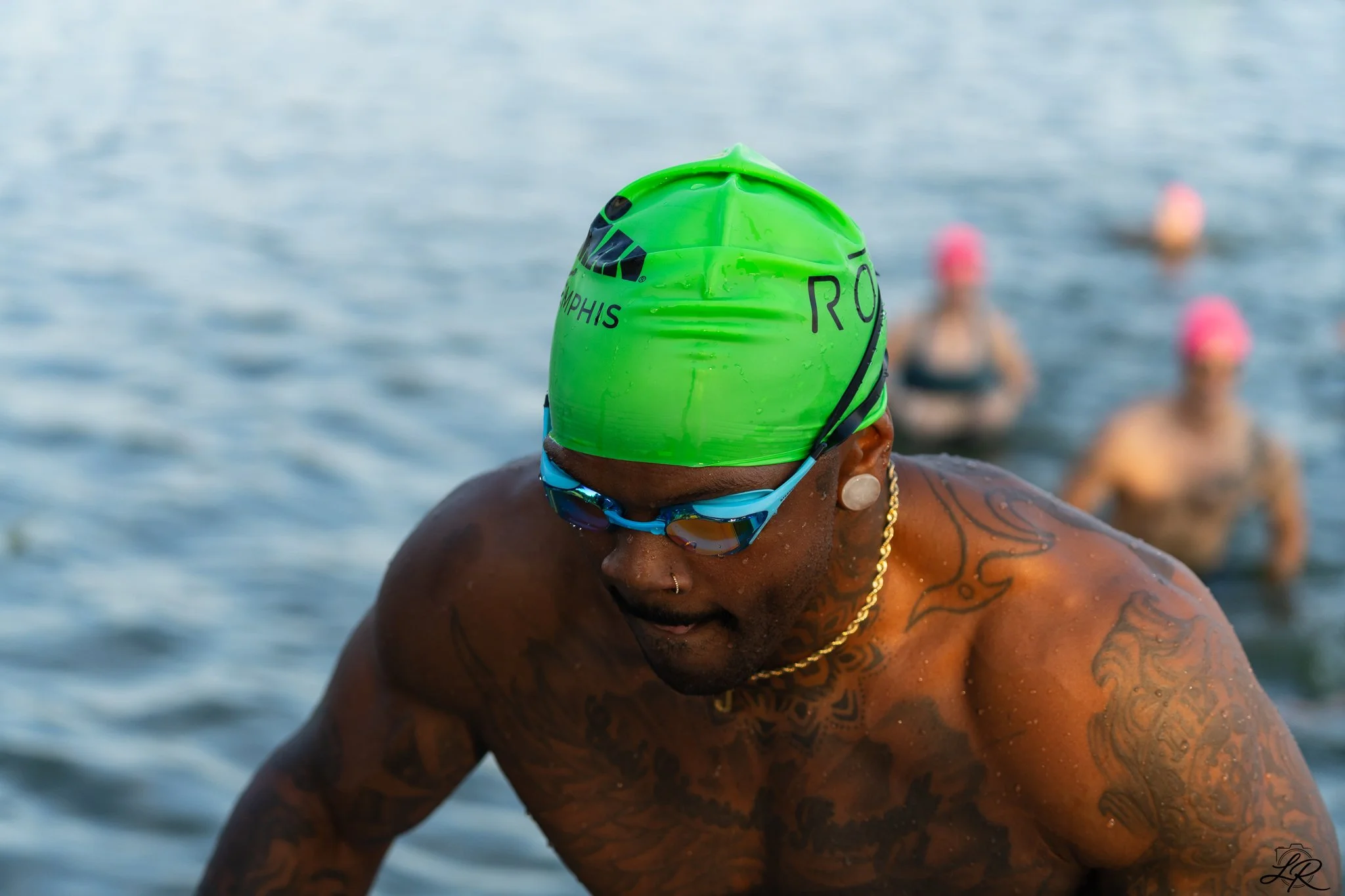A swimmer wearing a green swim cap with black text and goggles prepares to swim in a body of water, with other swimmers in the background wearing pink caps.