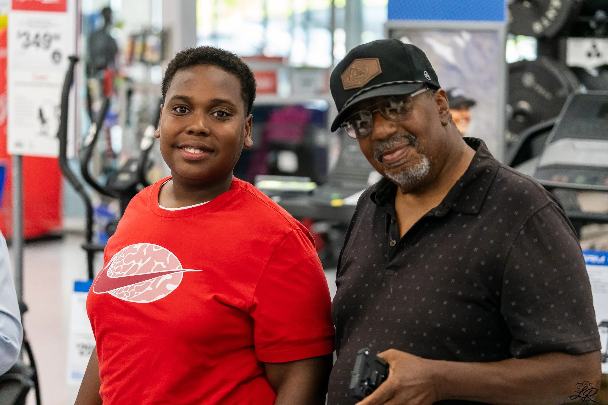 A young boy wearing a red Nike t-shirt with a brain diagram logo stands next to an older man wearing a black hat and glasses in a gym. They are smiling and appear to be looking at something off-camera.