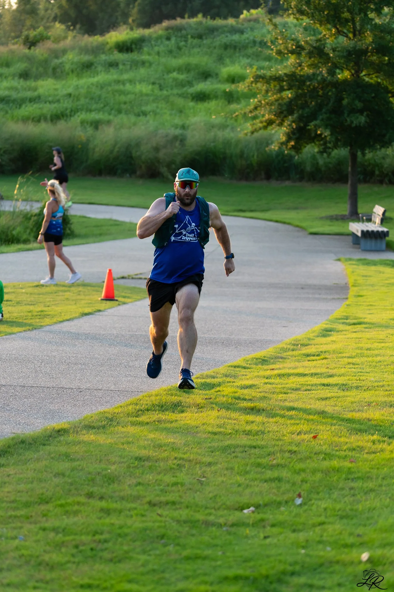 A man running on a paved trail in a park, wearing a blue tank top, black shorts, sunglasses, and a blue cap, with a backpack. Two women are in the background, one walking and the other running, surrounded by greenery and trees during the daytime.