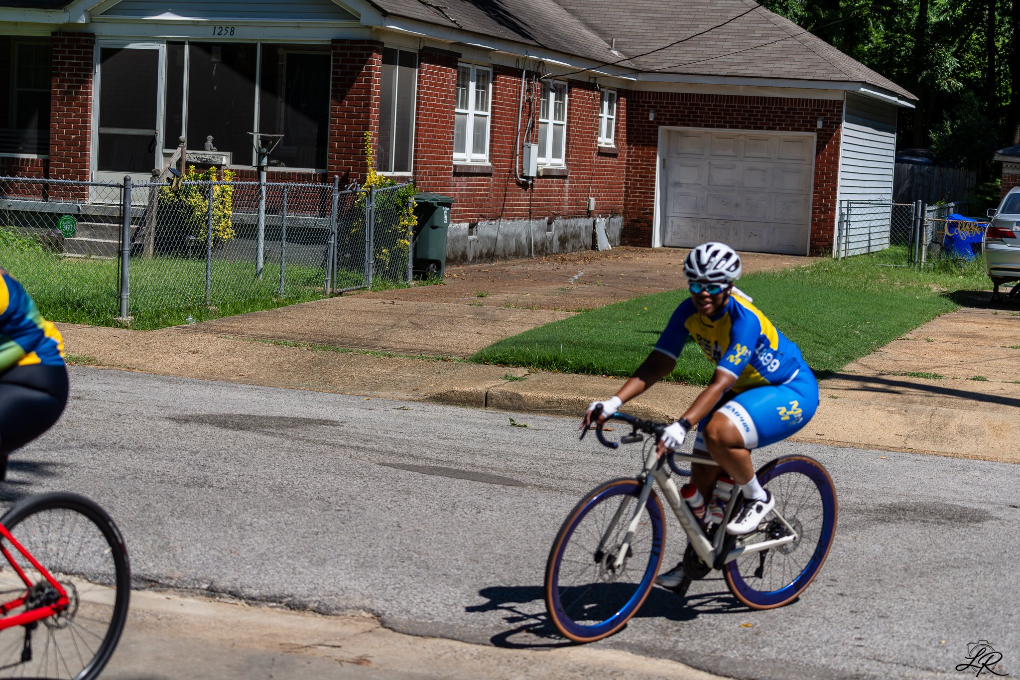 Cyclists riding on a residential street in front of a brick house with a garage, chain-link fence, and trees in the background.