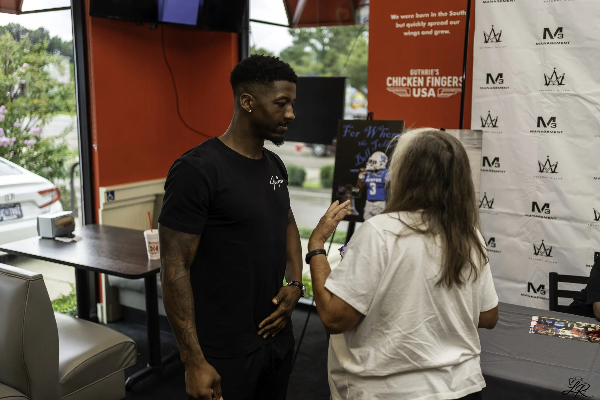 A young man with tattoos and earrings wearing a black t-shirt is talking to a woman with long hair and a white shirt at an indoor event. Behind them, there are banners, a counter, and a window showing outside greenery.