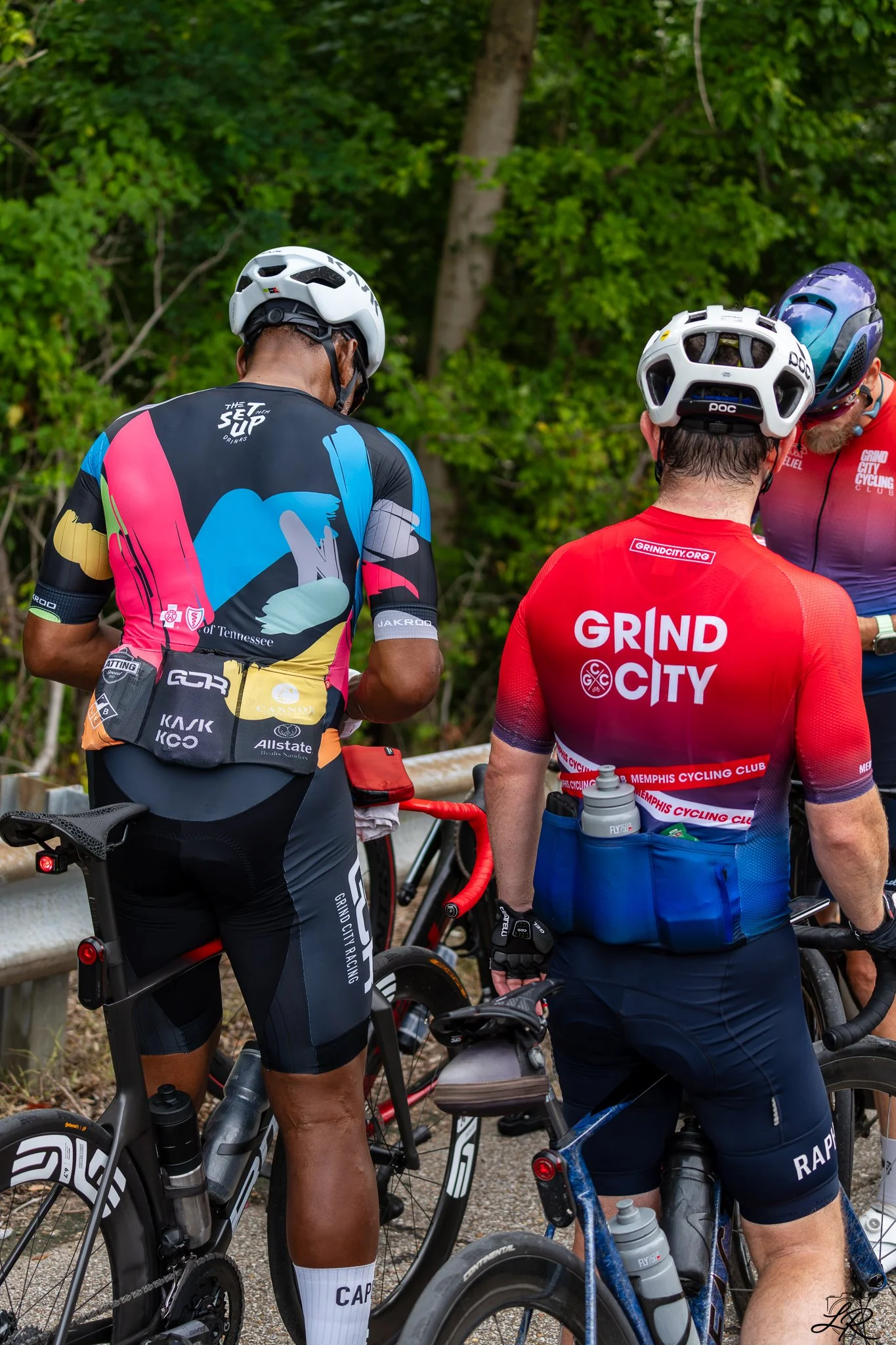 Three male cyclists wearing helmets and cycling gear stand together with their bikes on a wooded trail. Two have their backs to the camera, and one is on the right, partially visible, with green leafy trees in the background.