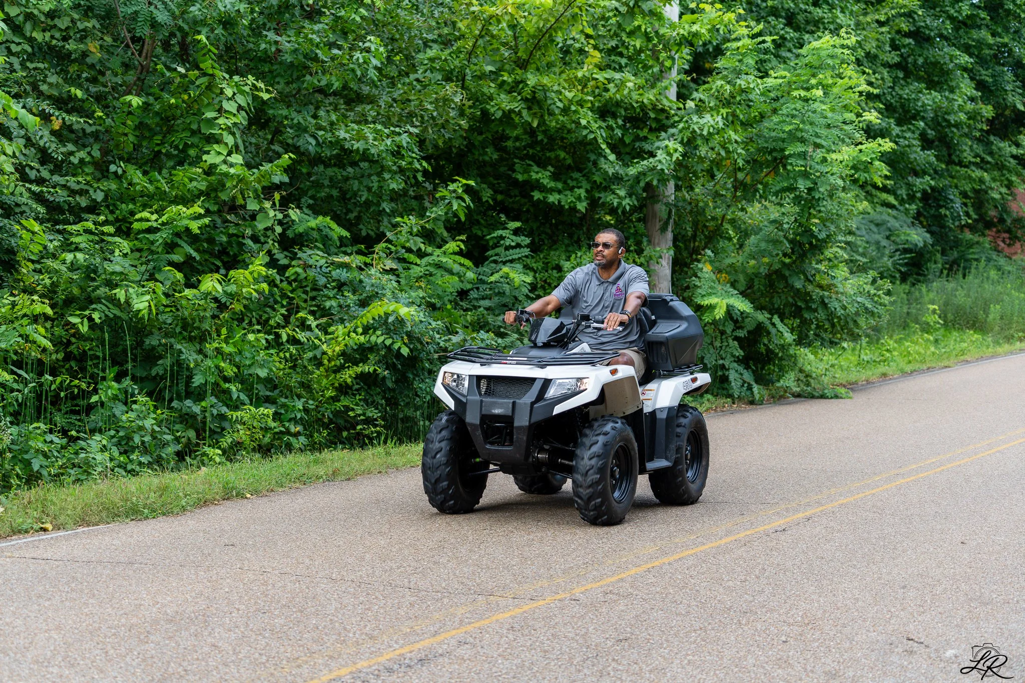 A man riding a black All-Terrain Vehicle (ATV) along a paved road next to dense green foliage and trees.