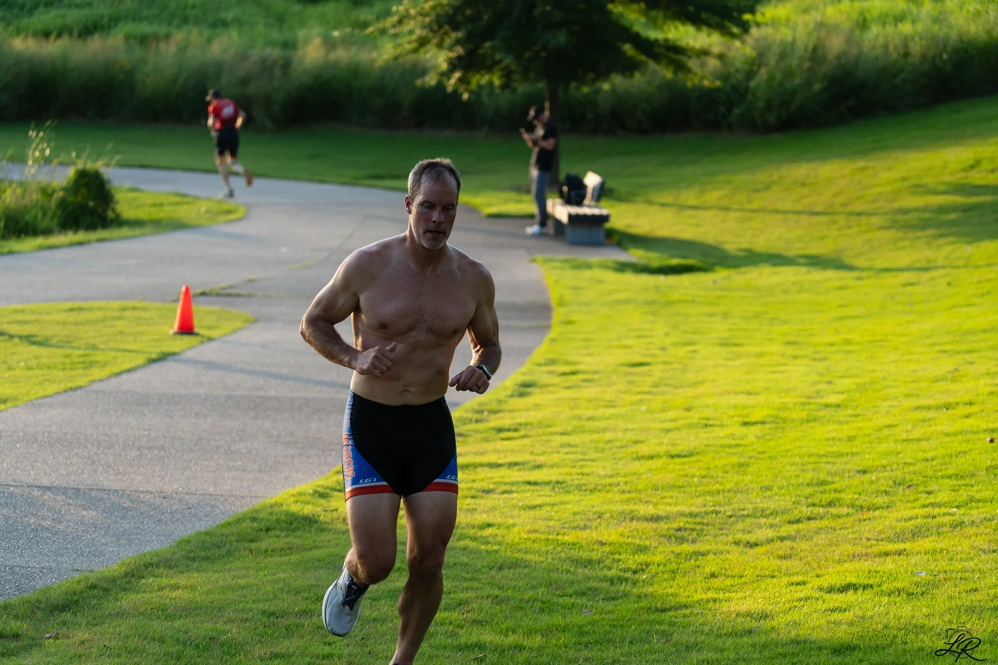 A shirtless man running on a paved path in a park during daytime, with two other people in the background and green grass and trees.