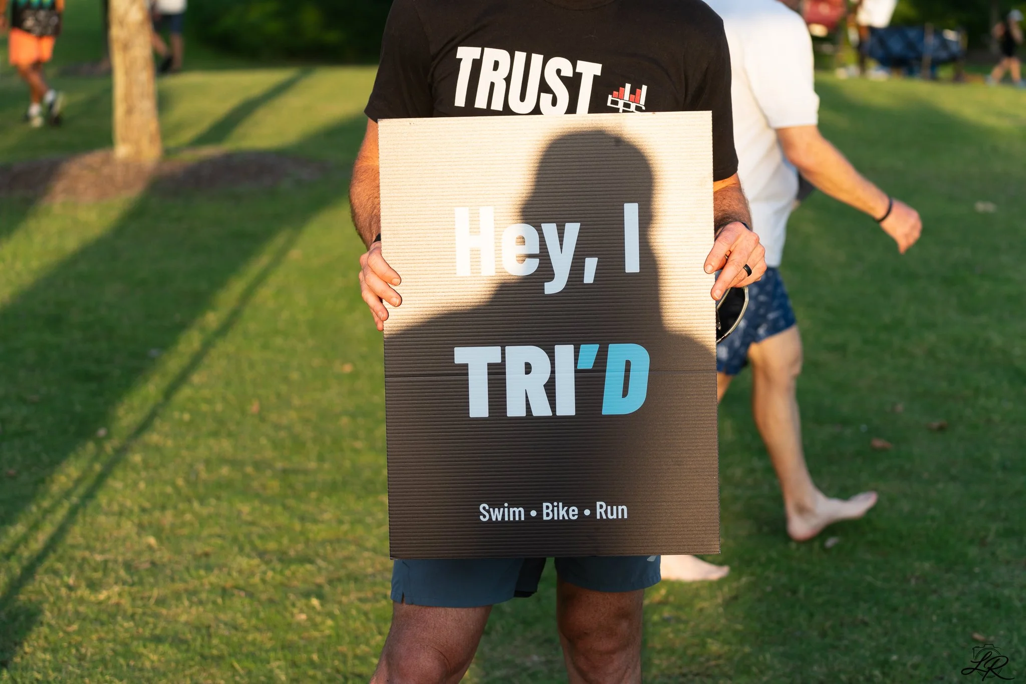 Person holding a sign that says 'Hey, I TRID' with smaller text 'Swim, Bike, Run' underneath, at an outdoor event or park during daytime.