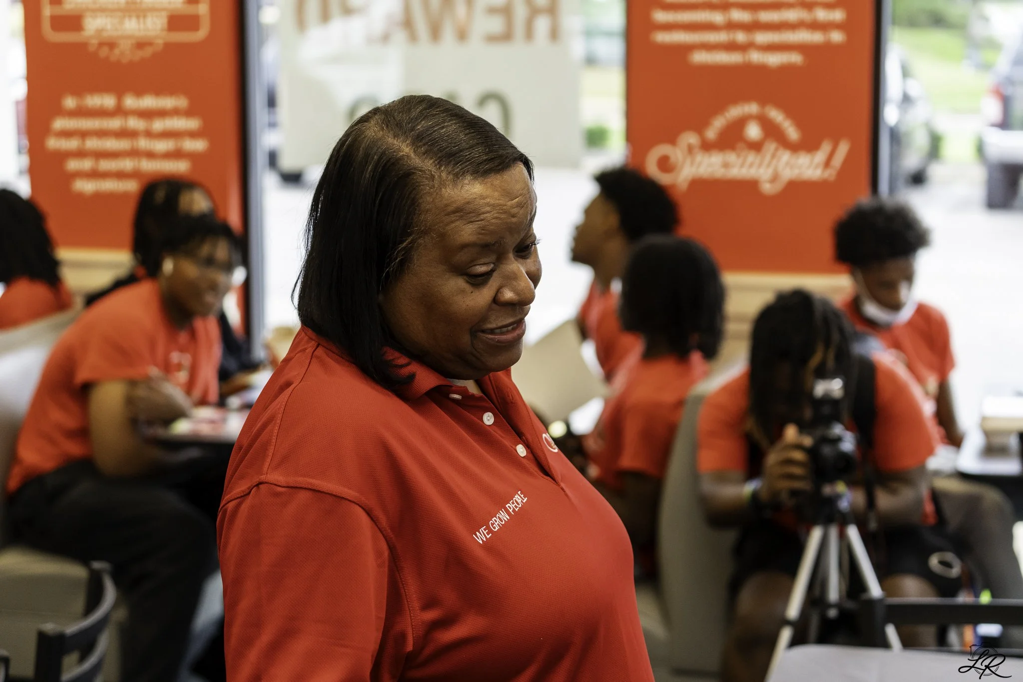 A woman in a red shirt with the words 'We Grow People' on it is looking downward at an event with other women in red shirts, some wearing glasses, and there are red banners in the background.