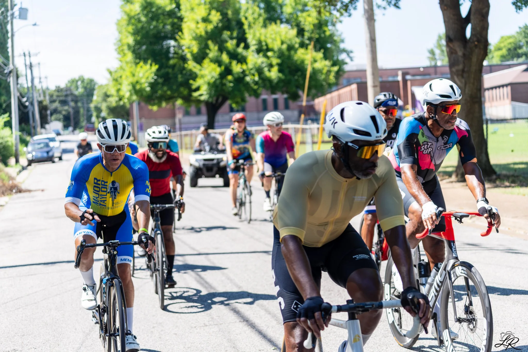 A group of cyclists participating in a race on a sunny street, some wearing helmets and sunglasses, with trees and buildings in the background.