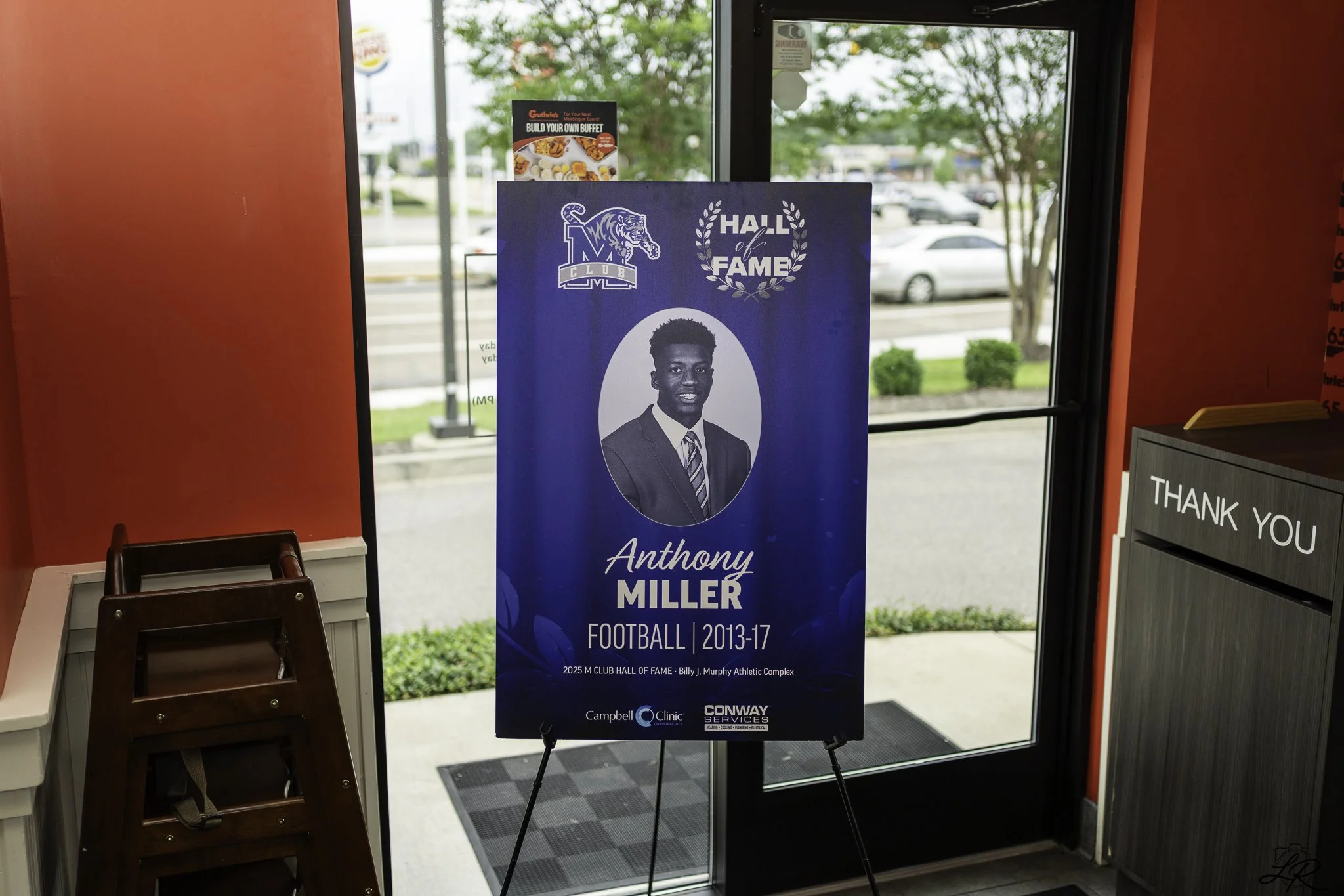 Indoor view of a restaurant or club entrance area with a large blue poster featuring a black and white portrait of Anthony Miller, a football player from 2013-2017, and logos for the Minnesota Vikings Hall of Fame and sponsors. There is a red wall on