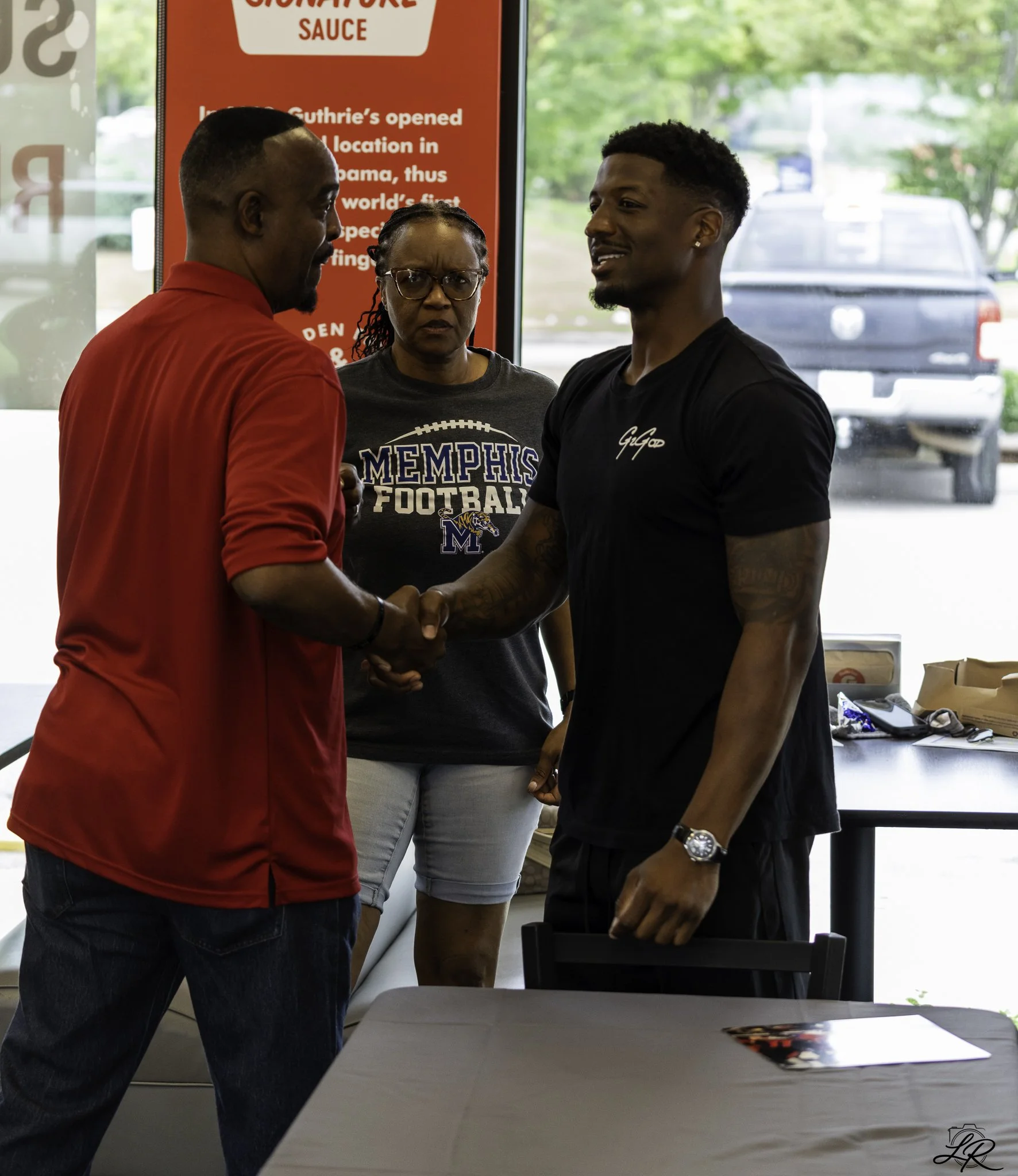 Two men shaking hands in a restaurant or diner, with a woman standing behind them and a red sign in the background, in front of a window showing a parking lot and trees outside.