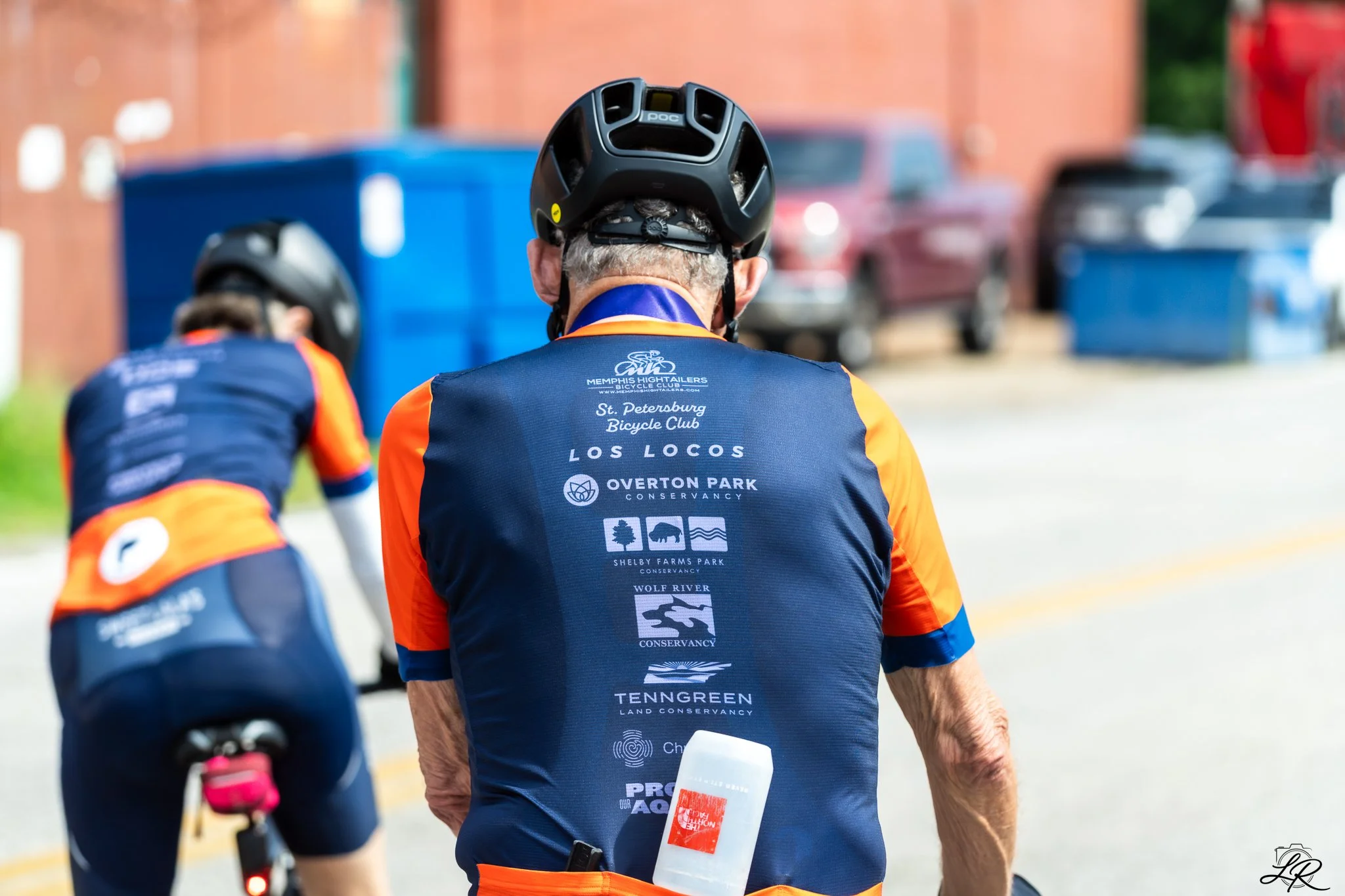 Back view of a cyclist wearing a black helmet and a navy and orange jersey with logos, riding outdoors on a paved path with parked cars and a brick building in the background.