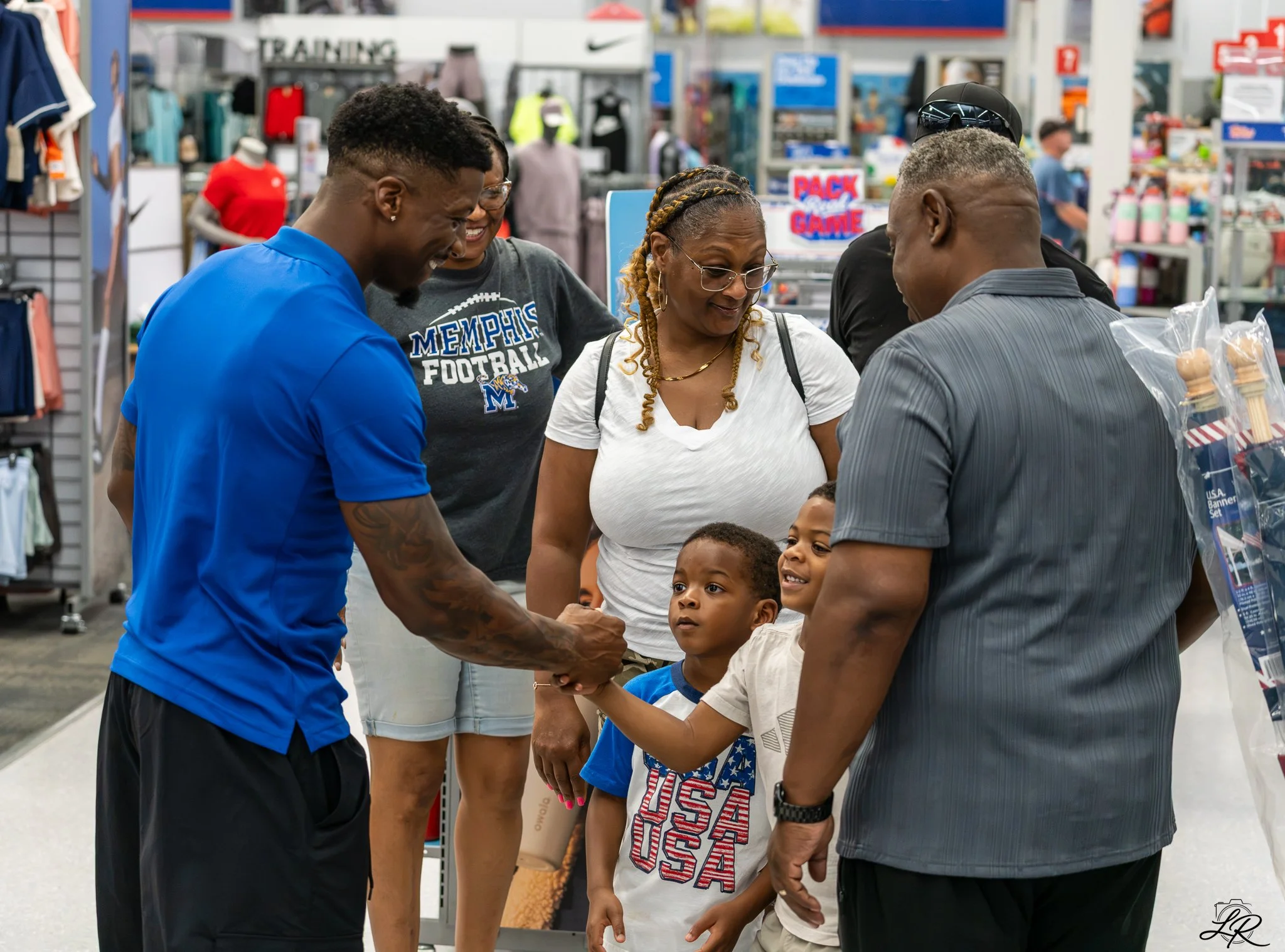 A group of African American people, including children and adults, are shopping in a sporting goods store. They are smiling, talking, and shaking hands, with sports apparel and accessories visible in the background.