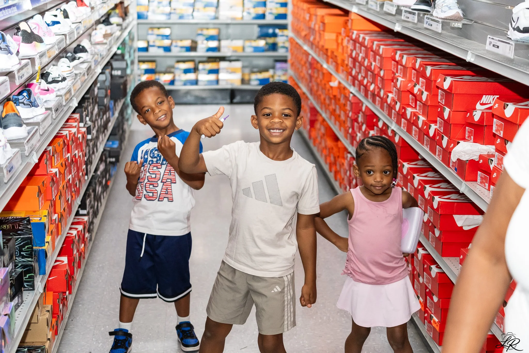 Three children standing in a shoe aisle of a store, smiling and posing for the camera. The store shelves are stocked with shoes and shoeboxes.