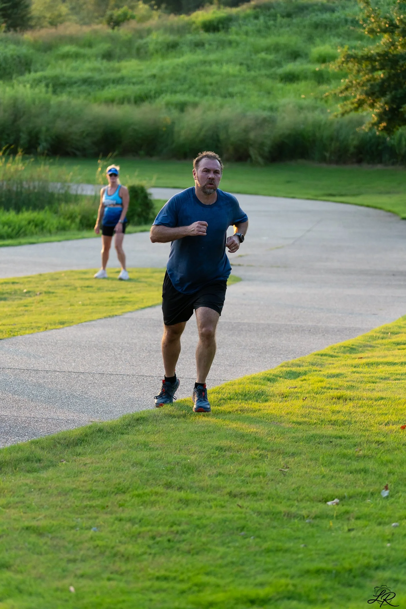 A man wearing a blue shirt and black shorts runs on a paved path in a grassy park, with a woman in a blue top and black shorts standing in the background. The park has lush green grass and trees, and the lighting suggests it is late afternoon or earl